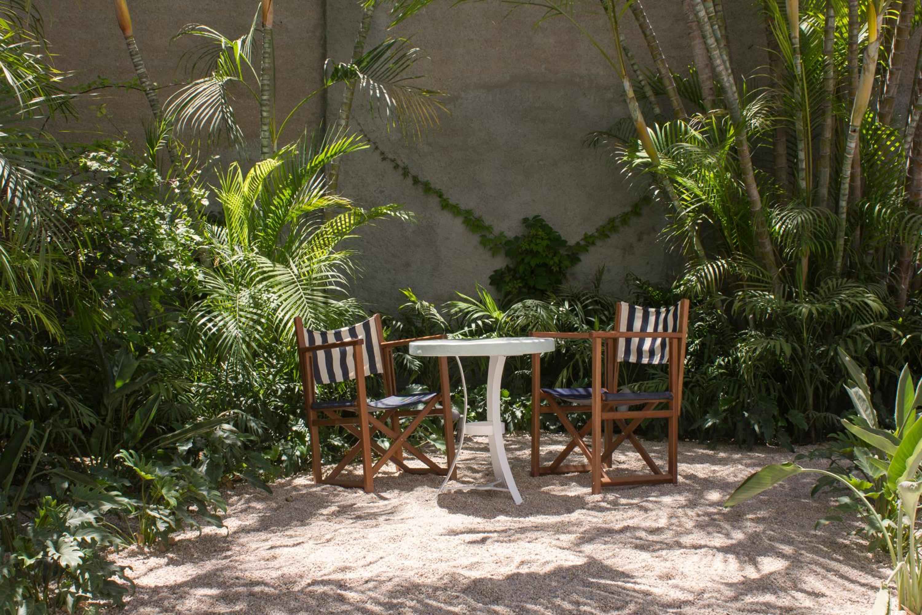 Dappled sunlight filters through swaying palms onto a secluded dining nook, where striped director's chairs invite intimate conversations beneath the desert garden's emerald canopy.