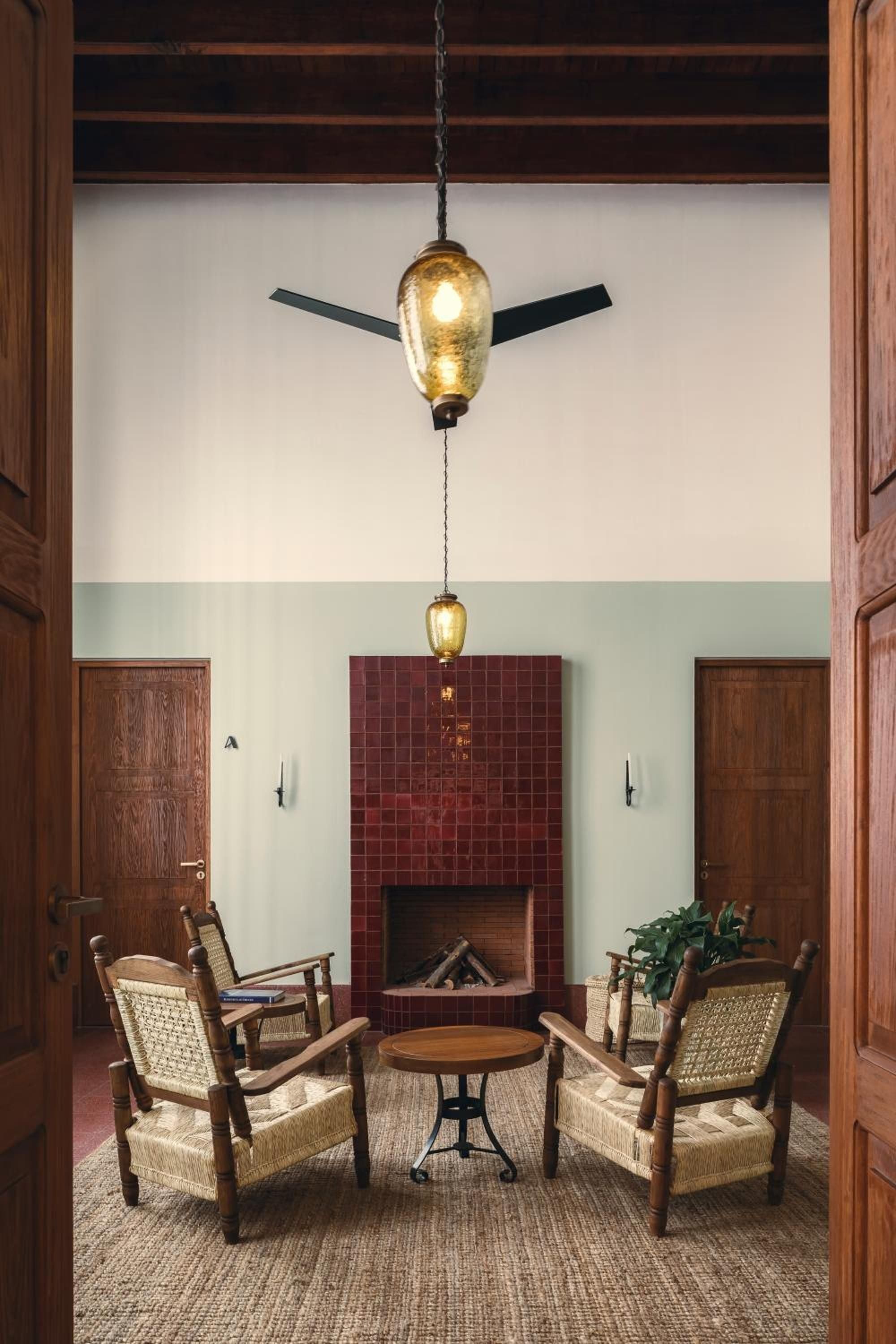 Woven cane chairs gather around a burnished fireplace beneath dramatic pendant lighting, where exposed beams and amber glass create intimate conversation nooks.