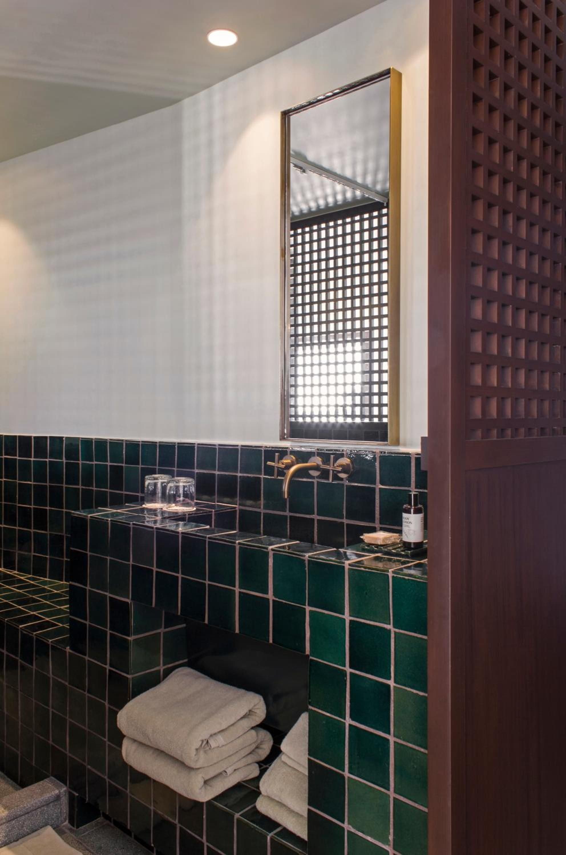 Emerald zellige tiles create striking geometric patterns in this thoughtfully designed bathroom, where brass fixtures and textured screens filter natural light into intimate, spa-like shadows.