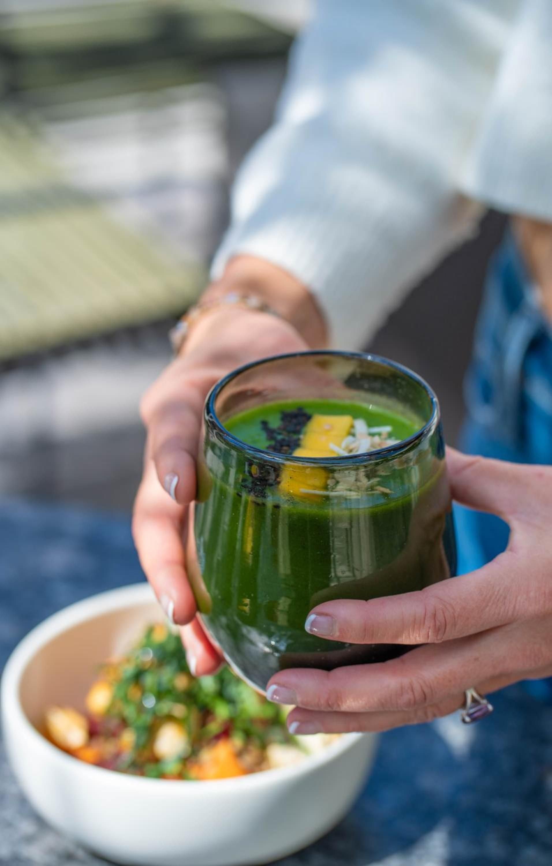 Hands cradle a vibrant green smoothie adorned with golden corn and fresh herbs, showcasing the hotel's commitment to locally-sourced, artfully presented cuisine.