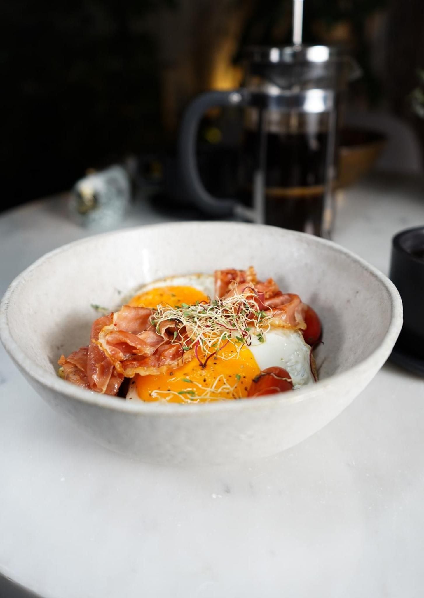 Farm-fresh eggs with silky yolks nestle alongside crispy bacon and delicate microgreens in a handcrafted ceramic bowl, embodying the hotel's commitment to locally-sourced breakfast artistry.