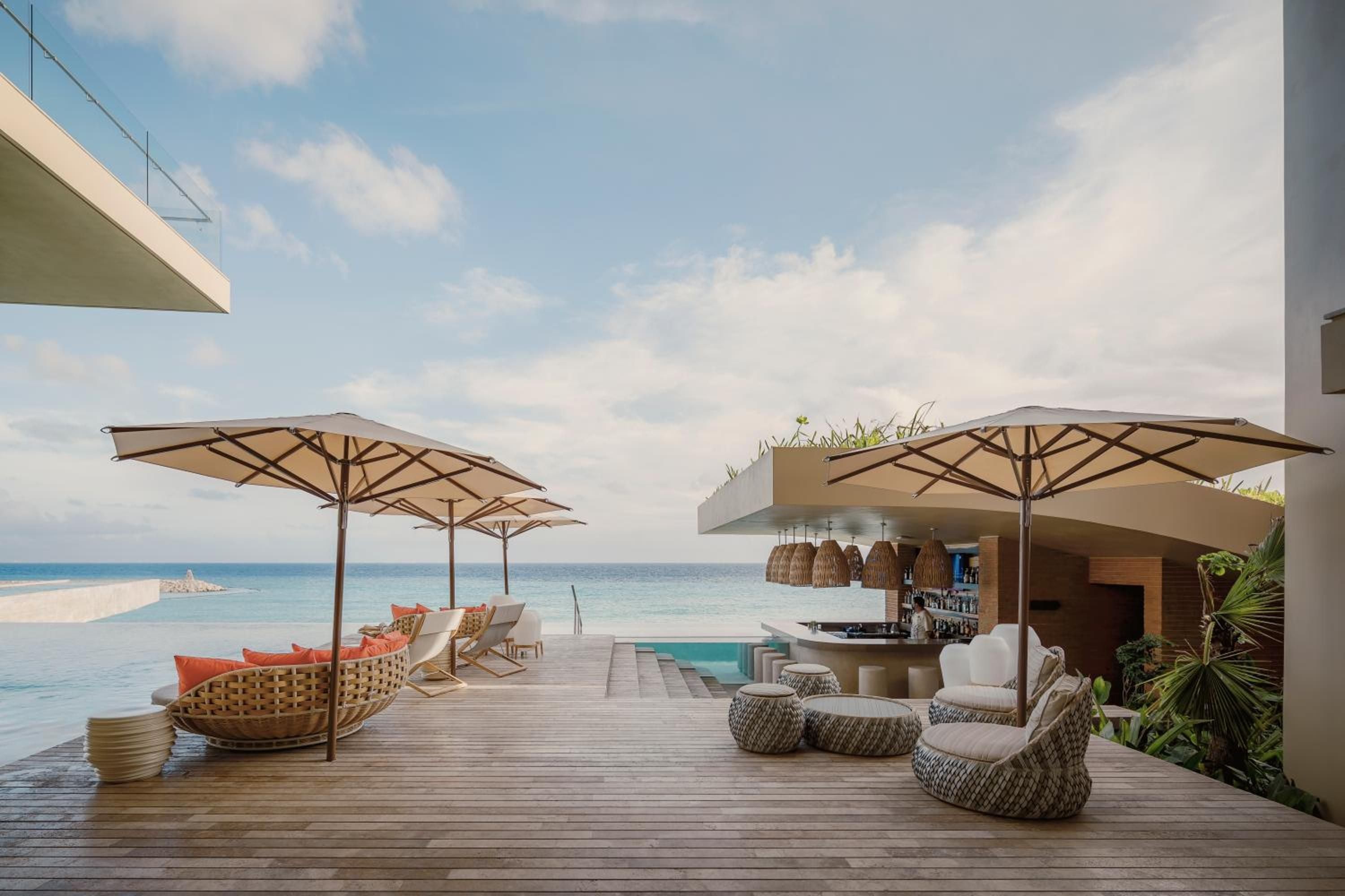 Woven rattan seating and pendant lights frame the beachfront bar, where turquoise waters meet honey-toned umbrellas on weathered teak decking.
