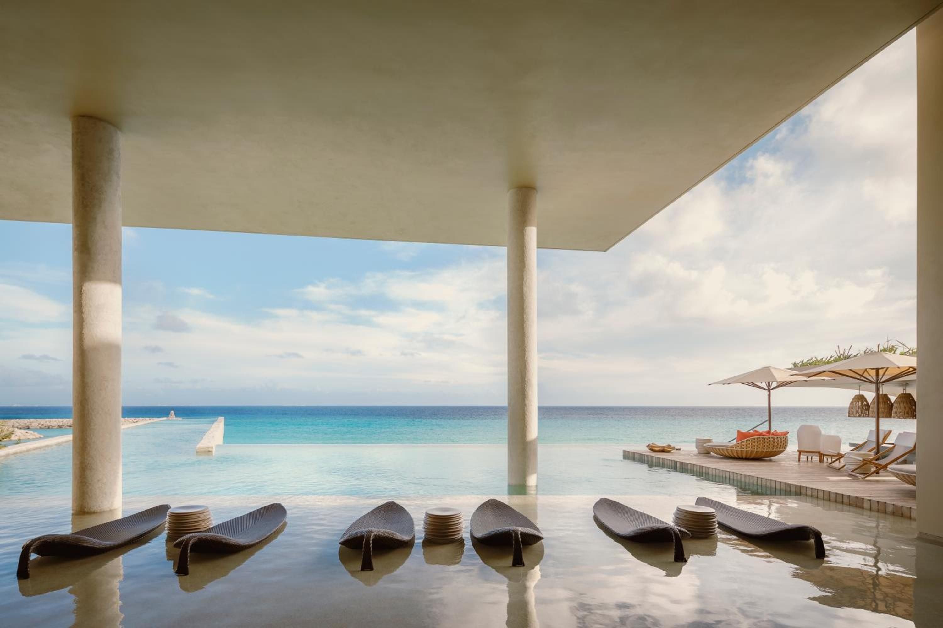 Infinity pool loungers appear to float on liquid glass, while the turquoise Caribbean stretches endlessly beyond minimalist columns framing this serene aquatic sanctuary.