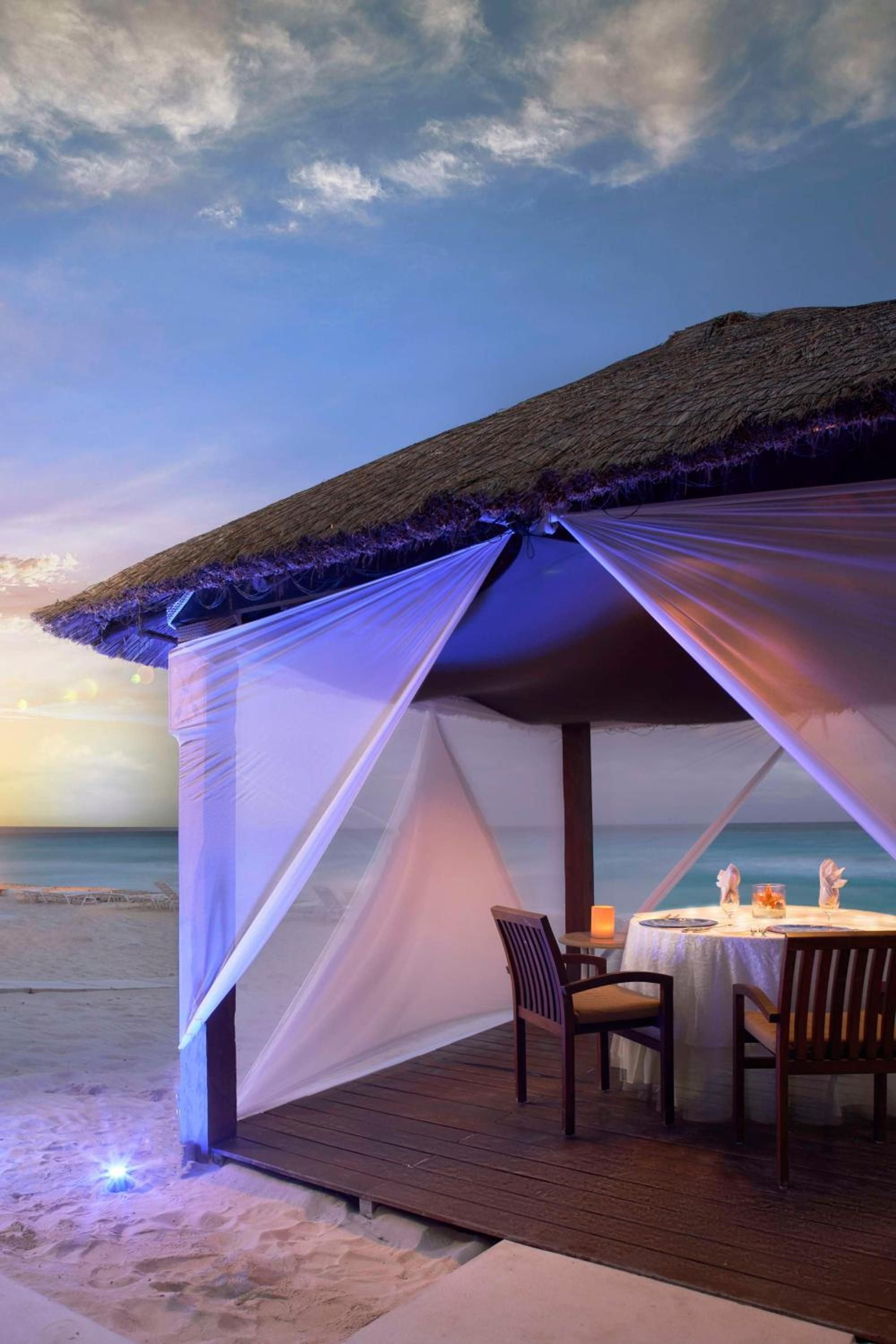 Purple twilight bathes an intimate beachside cabana where candlelit dinner awaits on warm sand, ocean waves whispering just steps away from your private thatched-roof sanctuary.