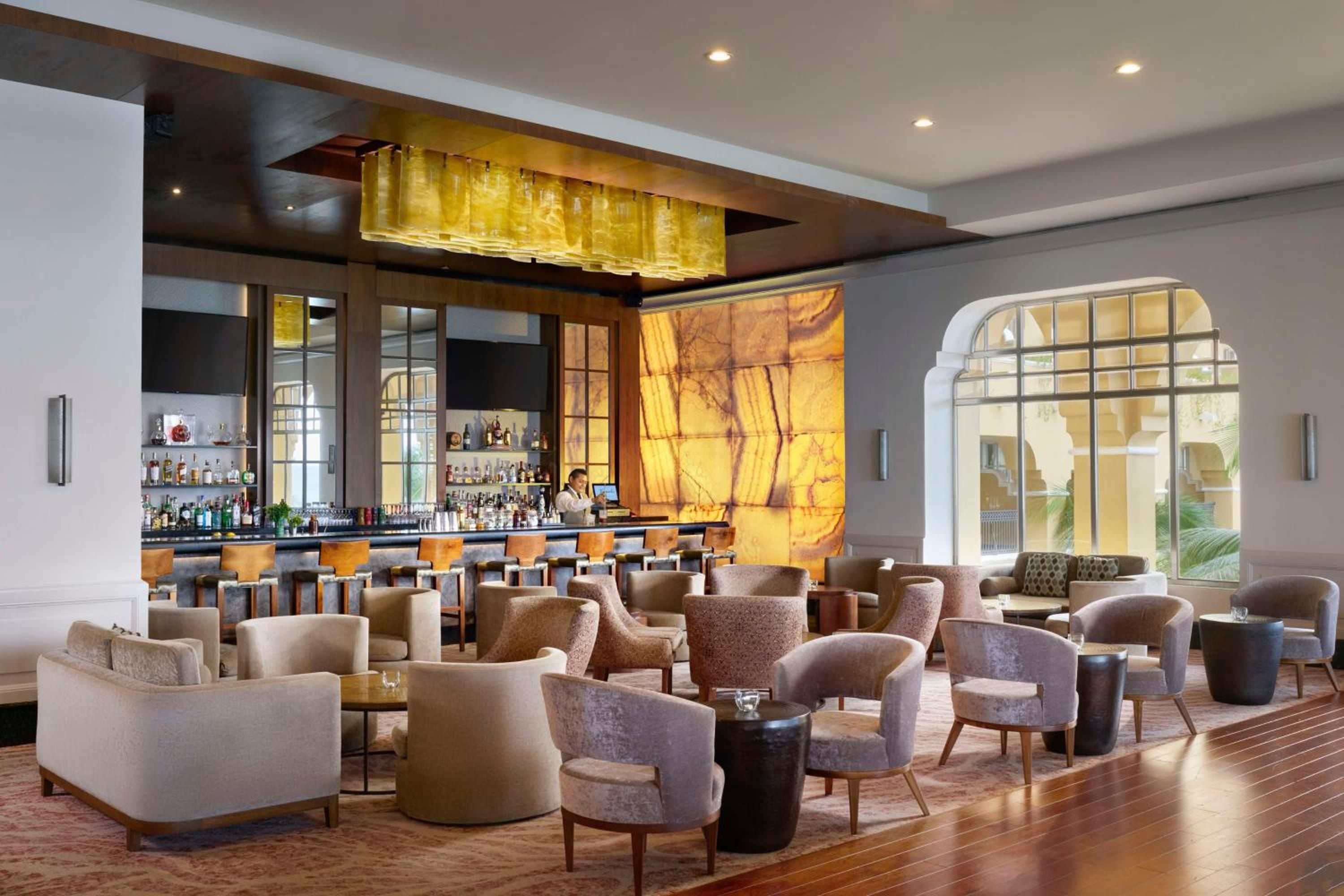 Warm amber lighting bathes plush armchairs and gleaming hardwood floors while bartenders craft cocktails beneath a stunning golden onyx feature wall.