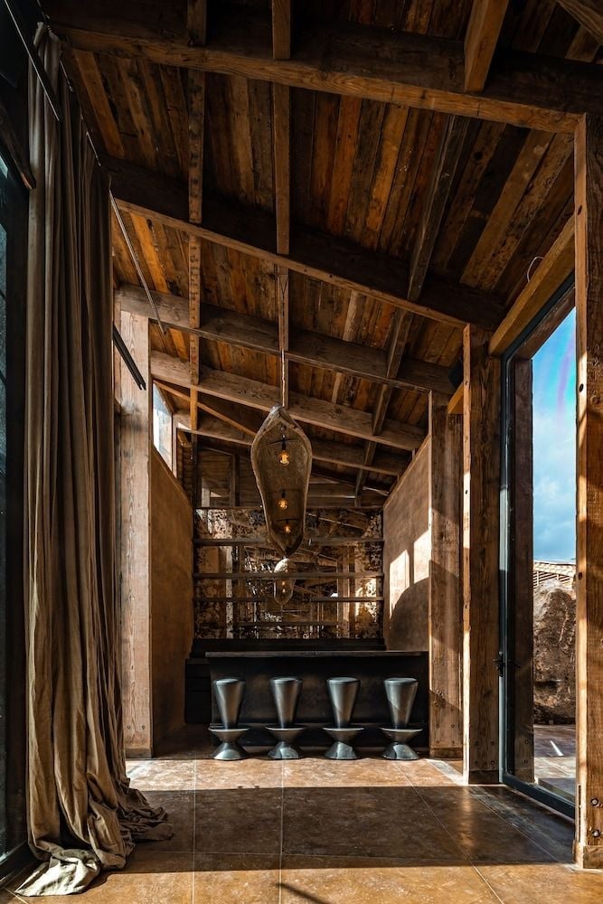 Exposed wooden beams frame a sophisticated tasting counter where four sleek stools await beneath rustic rafters, merging industrial distillery heritage with intimate hospitality.