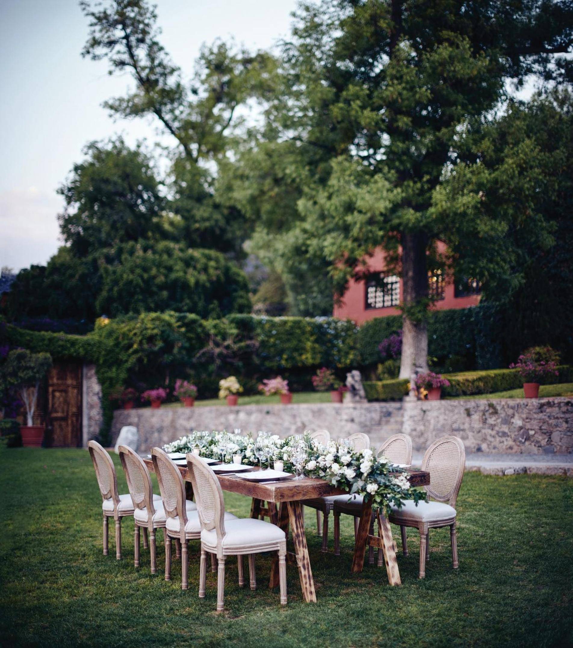 Candlelit dining unfolds on manicured lawns beneath towering trees, where Louis XVI chairs and fresh florals create an enchanting al fresco tableau against colonial pink walls.