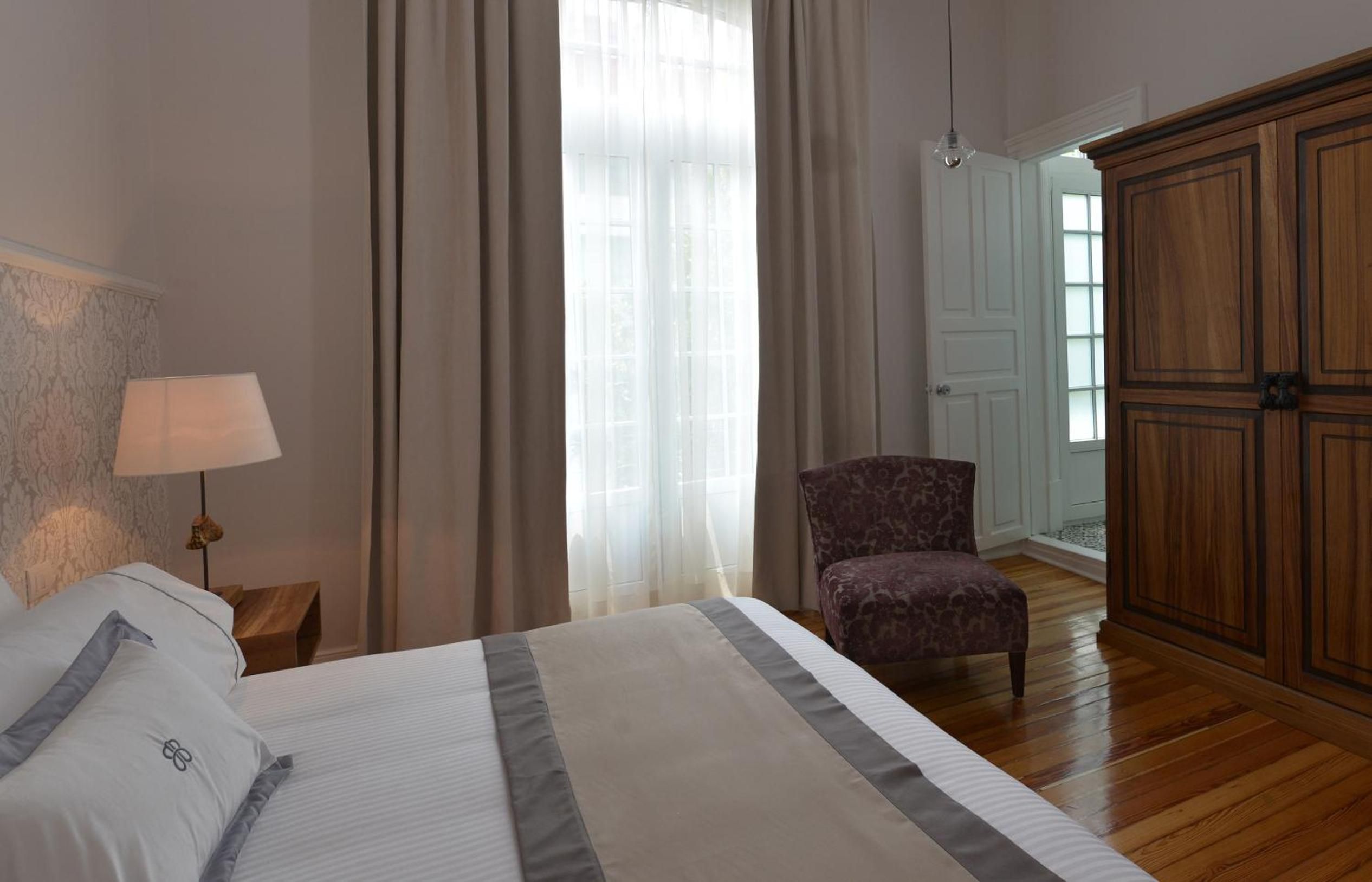Warm hardwood floors and rich wooden armoire create an intimate sanctuary, where soft natural light filters through taupe curtains onto crisp linens and burgundy velvet seating.