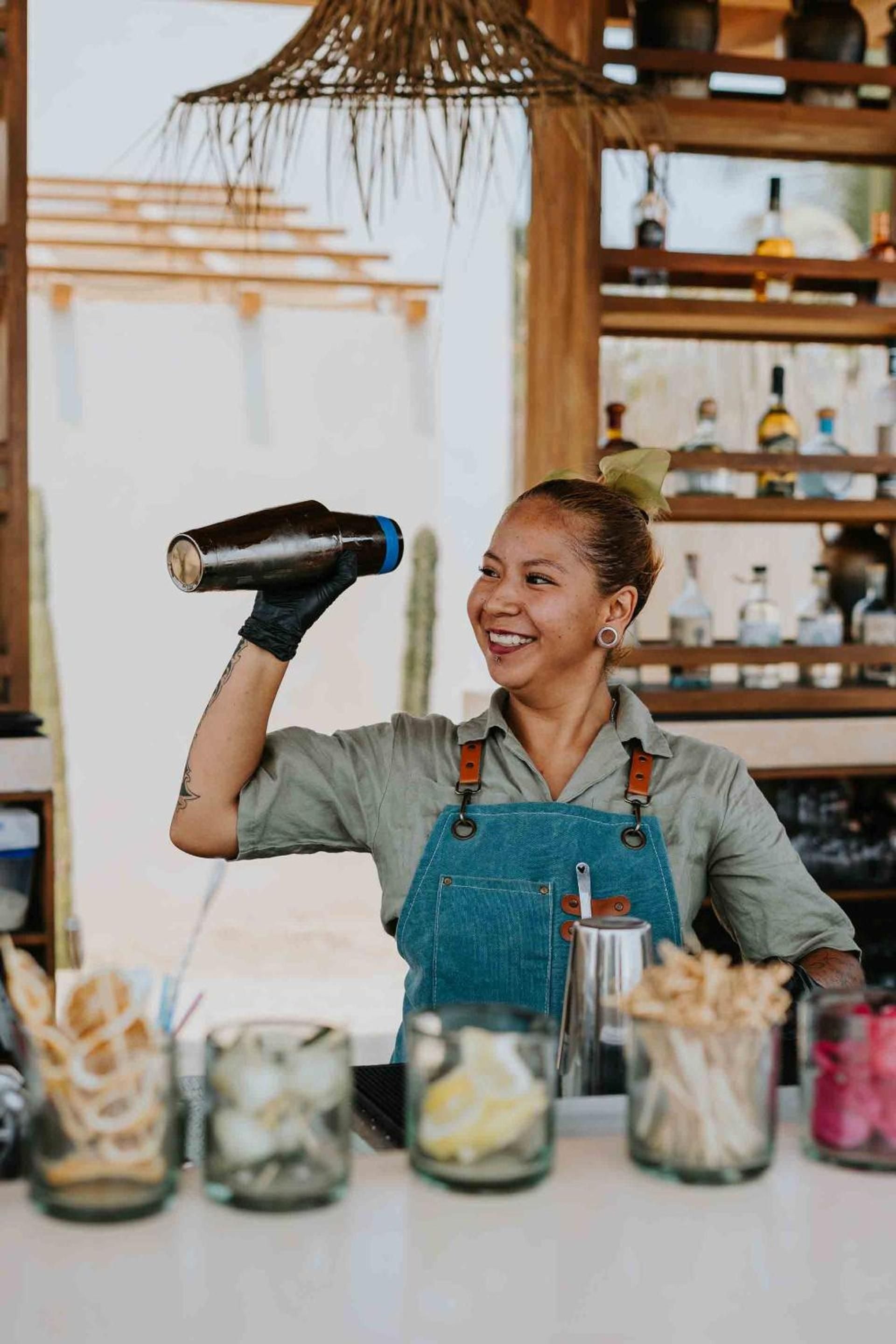 Behind Casa Yuma's bar, skilled bartenders craft mezcal cocktails with infectious enthusiasm, their tools gleaming under woven lampshades in the sun-drenched space.