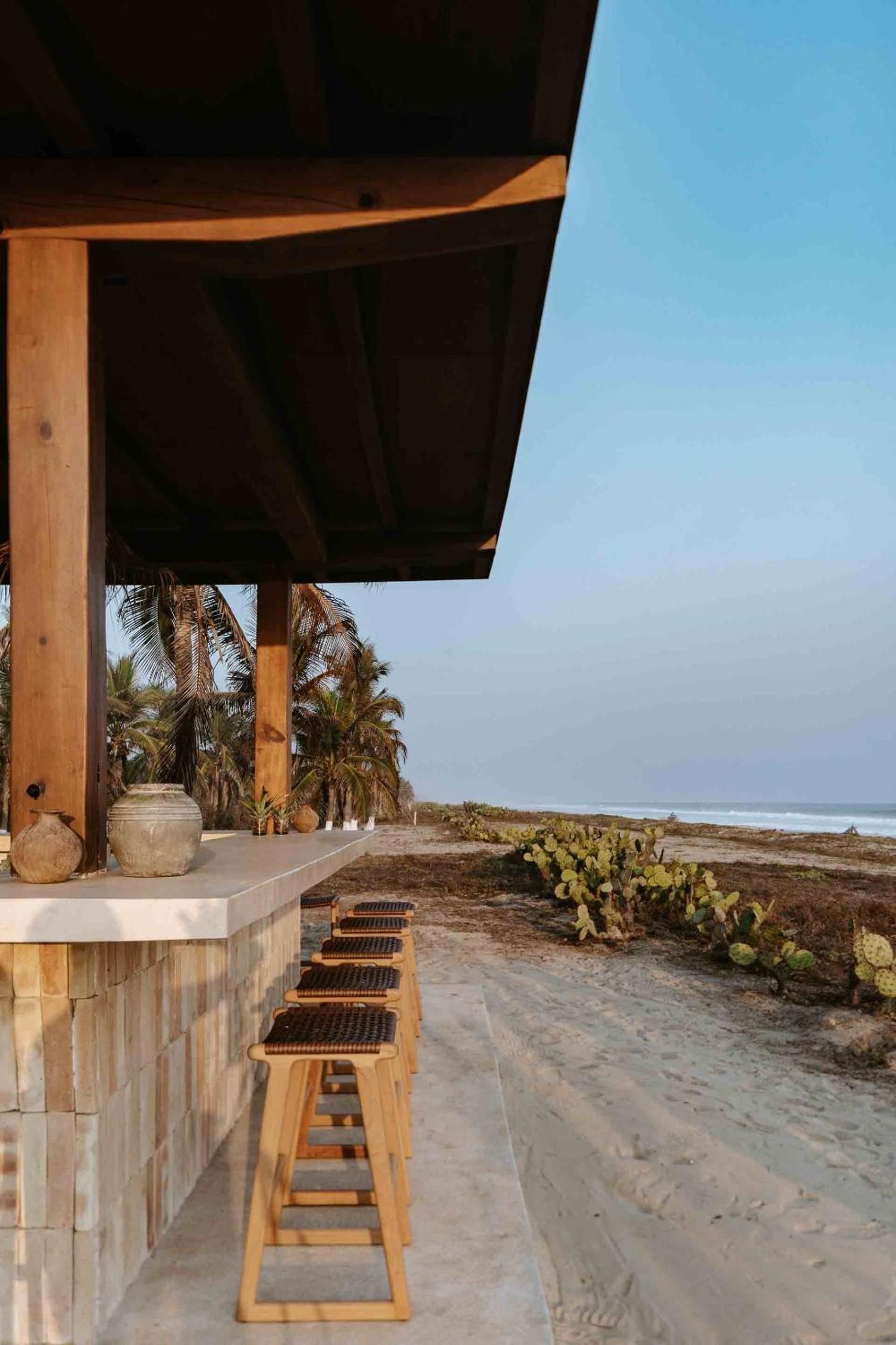 Woven bar stools line the outdoor counter where ceramic vessels catch golden hour light, while palm trees frame the wild Pacific coastline beyond.