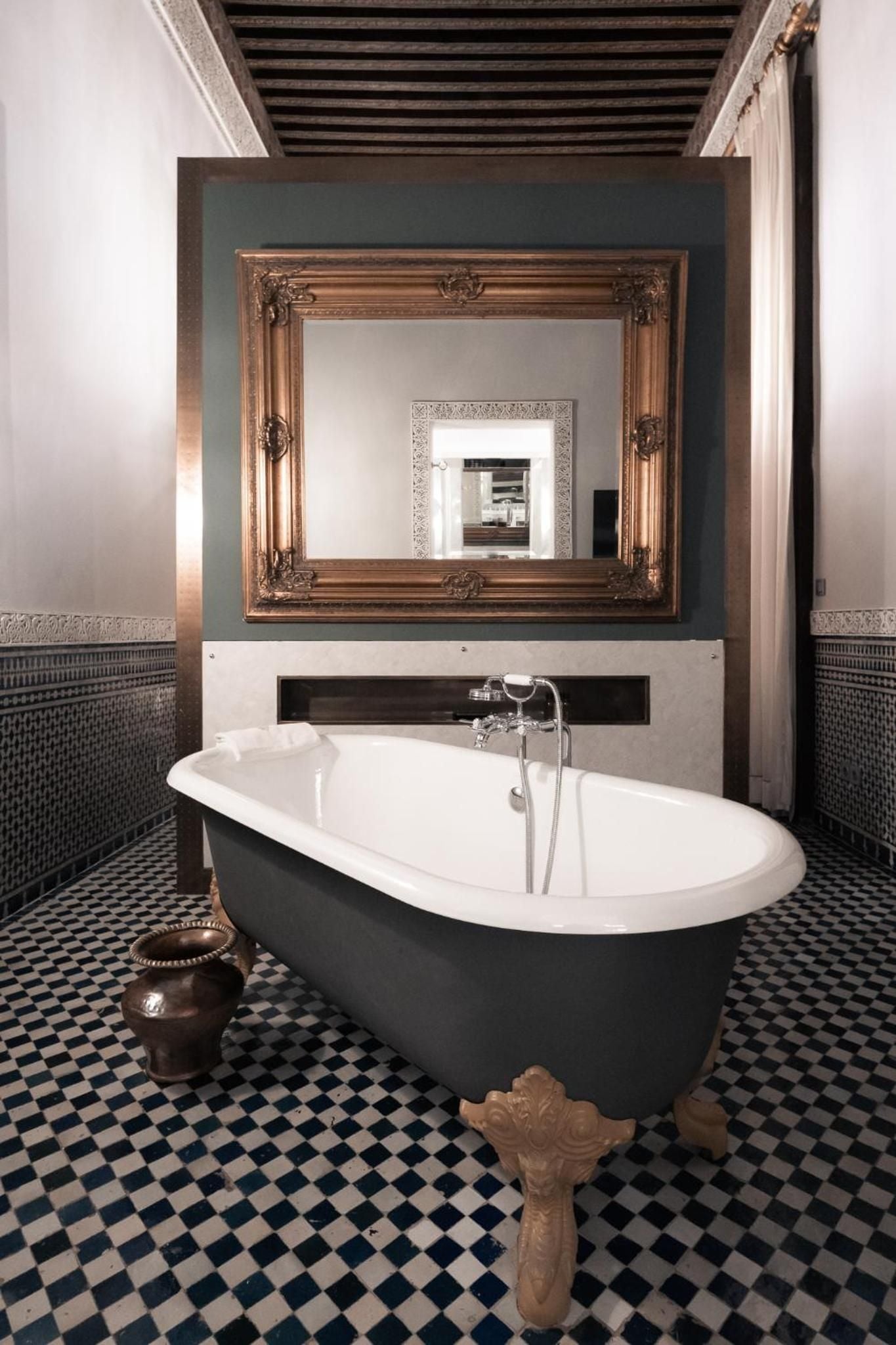 Ornate golden mirrors and geometric zellige tiles frame a sculptural clawfoot tub, where Moroccan craftsmanship meets luxurious indulgence beneath exposed cedar beams.