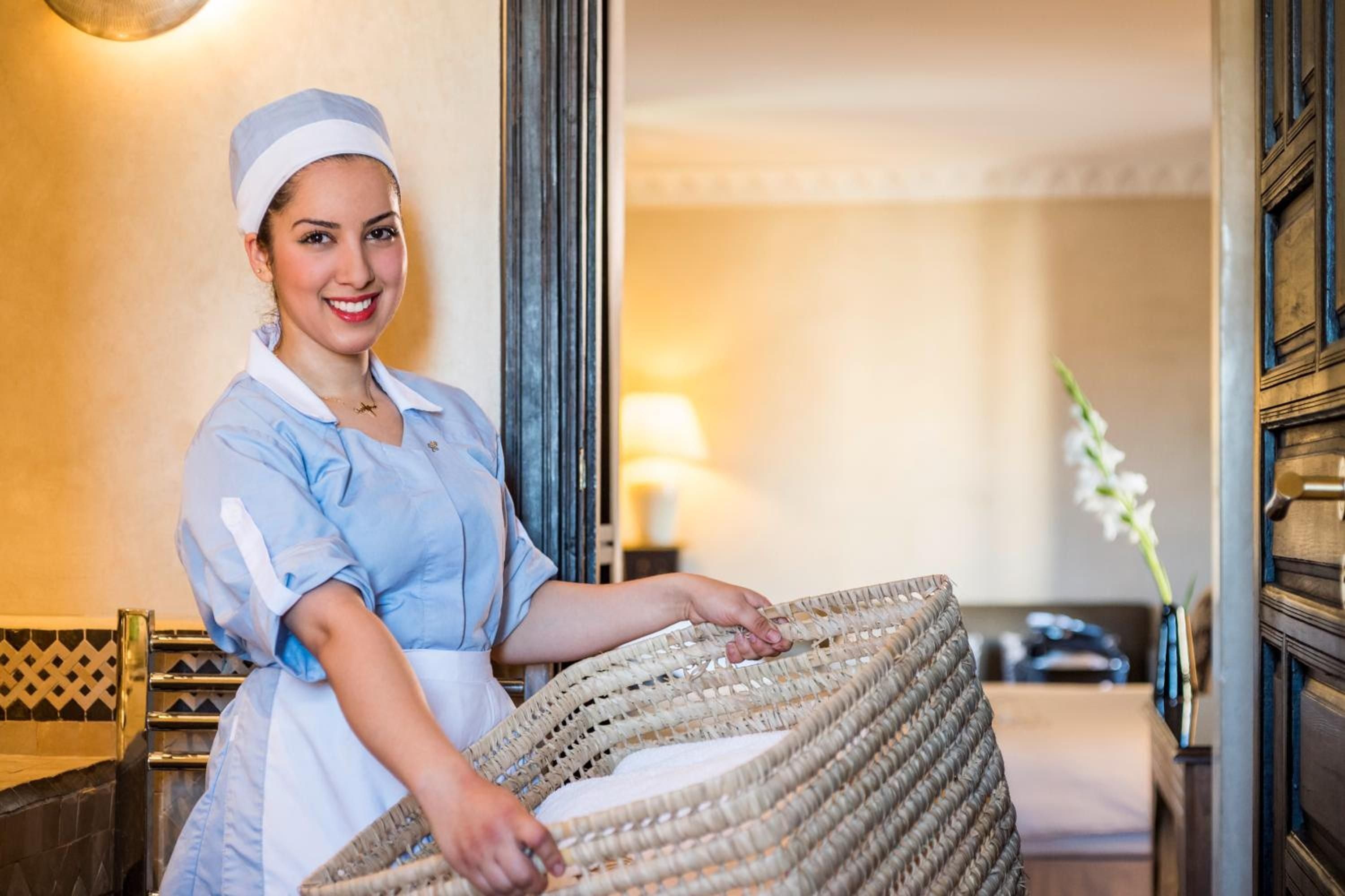 Staff in traditional blue uniforms prepare woven baskets in sunlit corridors, where Moroccan craftsmanship meets attentive service beneath warm amber lighting.