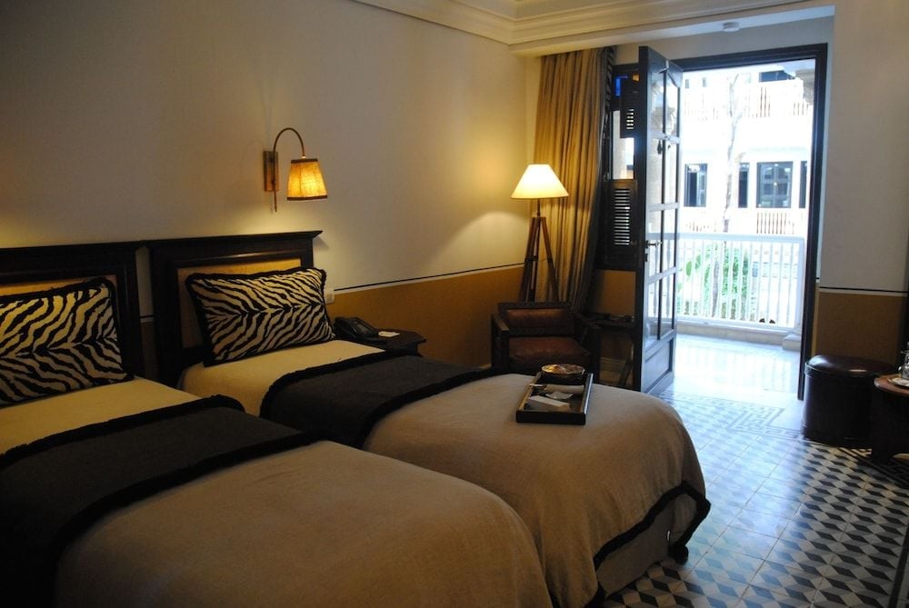 Twin beds adorned with zebra-print pillows invite relaxation, while warm lamplight and an open balcony door reveal glimpses of Essaouira's historic medina beyond.