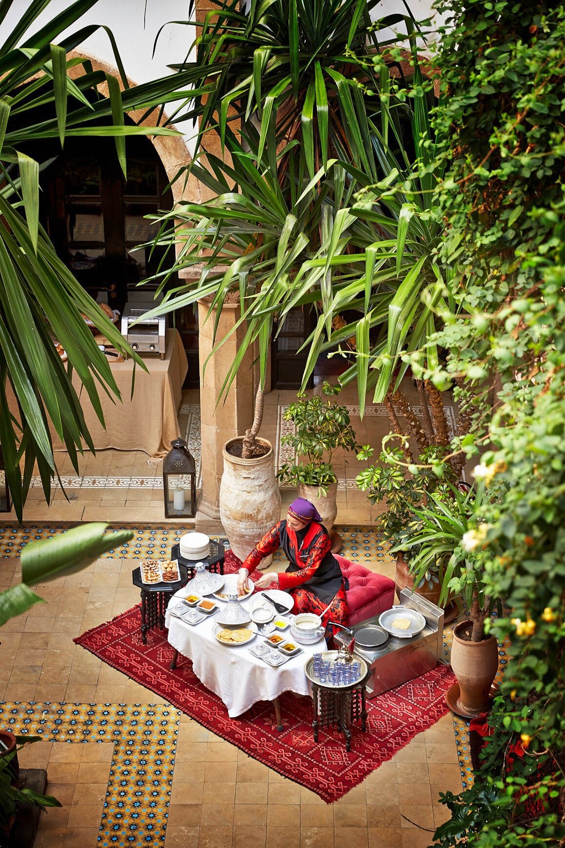 Beneath towering palms and climbing vines, traditional Moroccan breakfast unfolds on handwoven carpets in this enchanting riad courtyard, where geometric tiles frame moments of pure serenity.