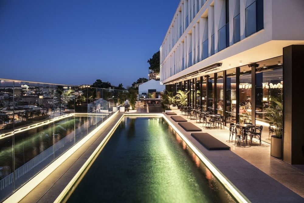 Golden hour transforms the rooftop pool into a luminous ribbon, while the open-air restaurant buzzes with energy against Lisbon's twinkling cityscape below.