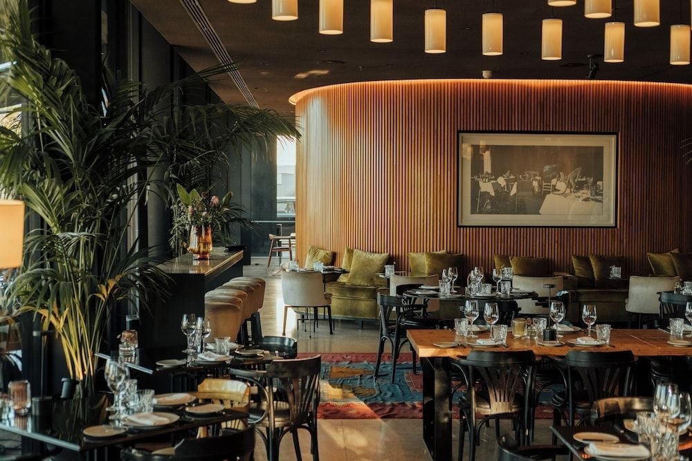 Warm pendant lights cascade over intimate dining tables while vertical wood slats create a sophisticated backdrop, blending modern design with inviting comfort in the hotel's signature restaurant.