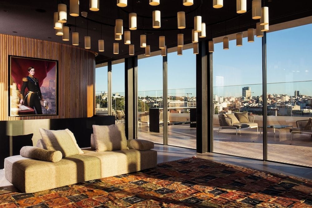 Floor-to-ceiling windows frame Lisbon's rooftops from this sumptuous suite, where sculptural pendant lights dance above plush seating and rich Persian rugs warm polished floors.