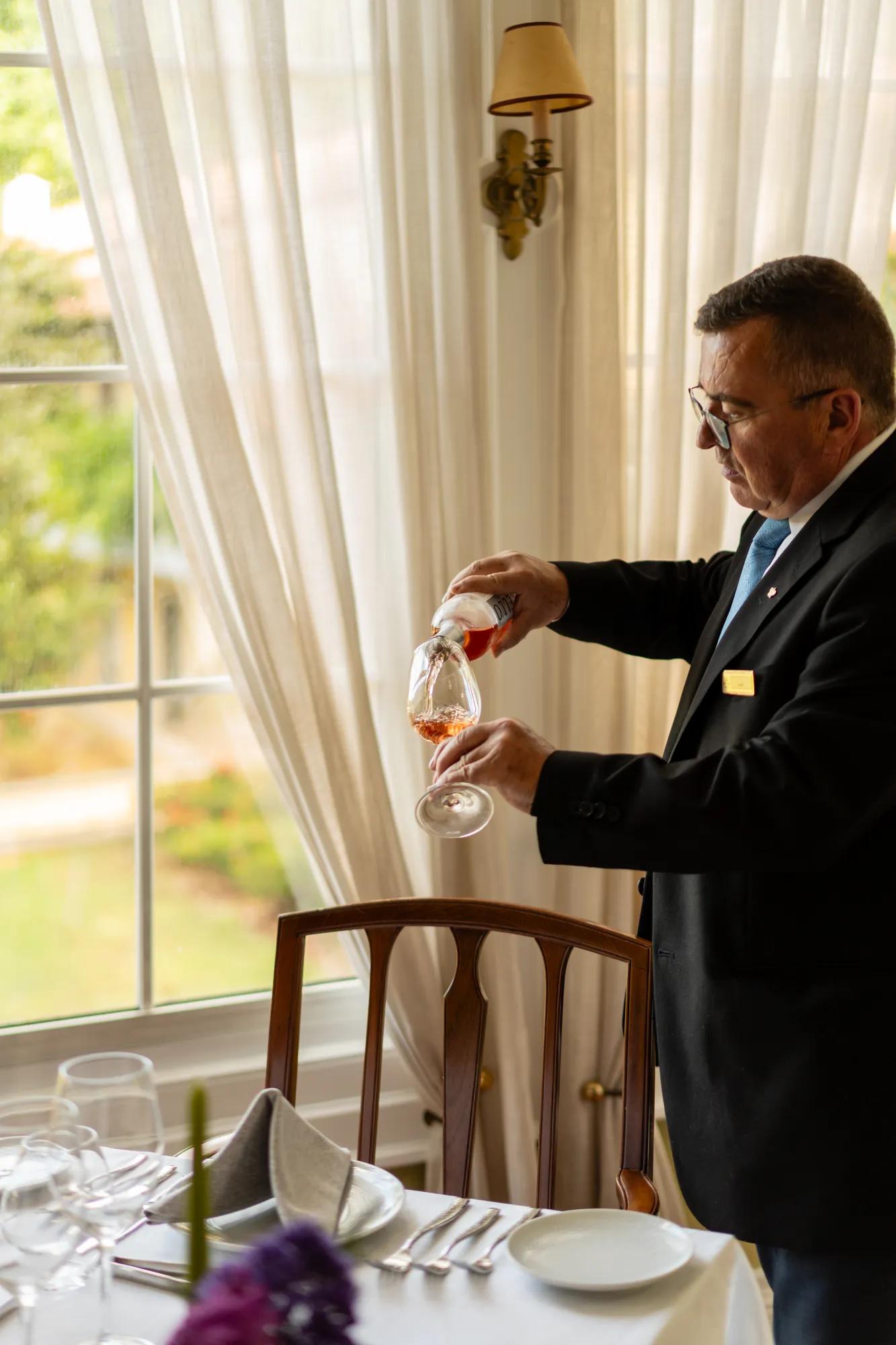 A sommelier's practiced hands pour amber wine beside sun-drenched windows, where crisp linens and purple blooms create an intimate dining tableau.