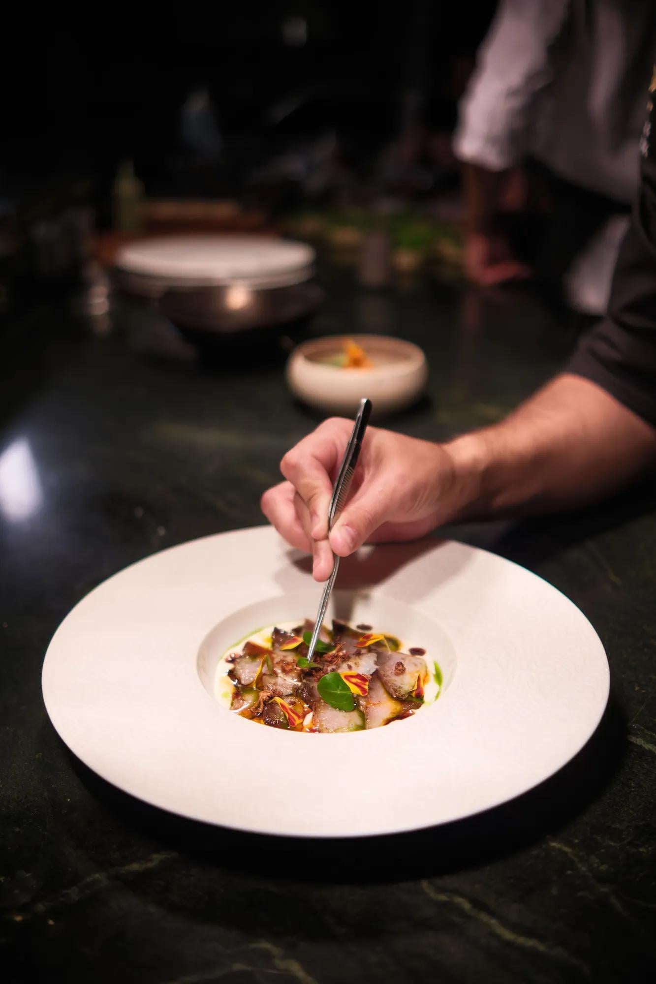 Precision meets artistry as a chef adds finishing touches to an elegantly plated dish, each microgreen carefully positioned in the intimate dining room's warm glow.