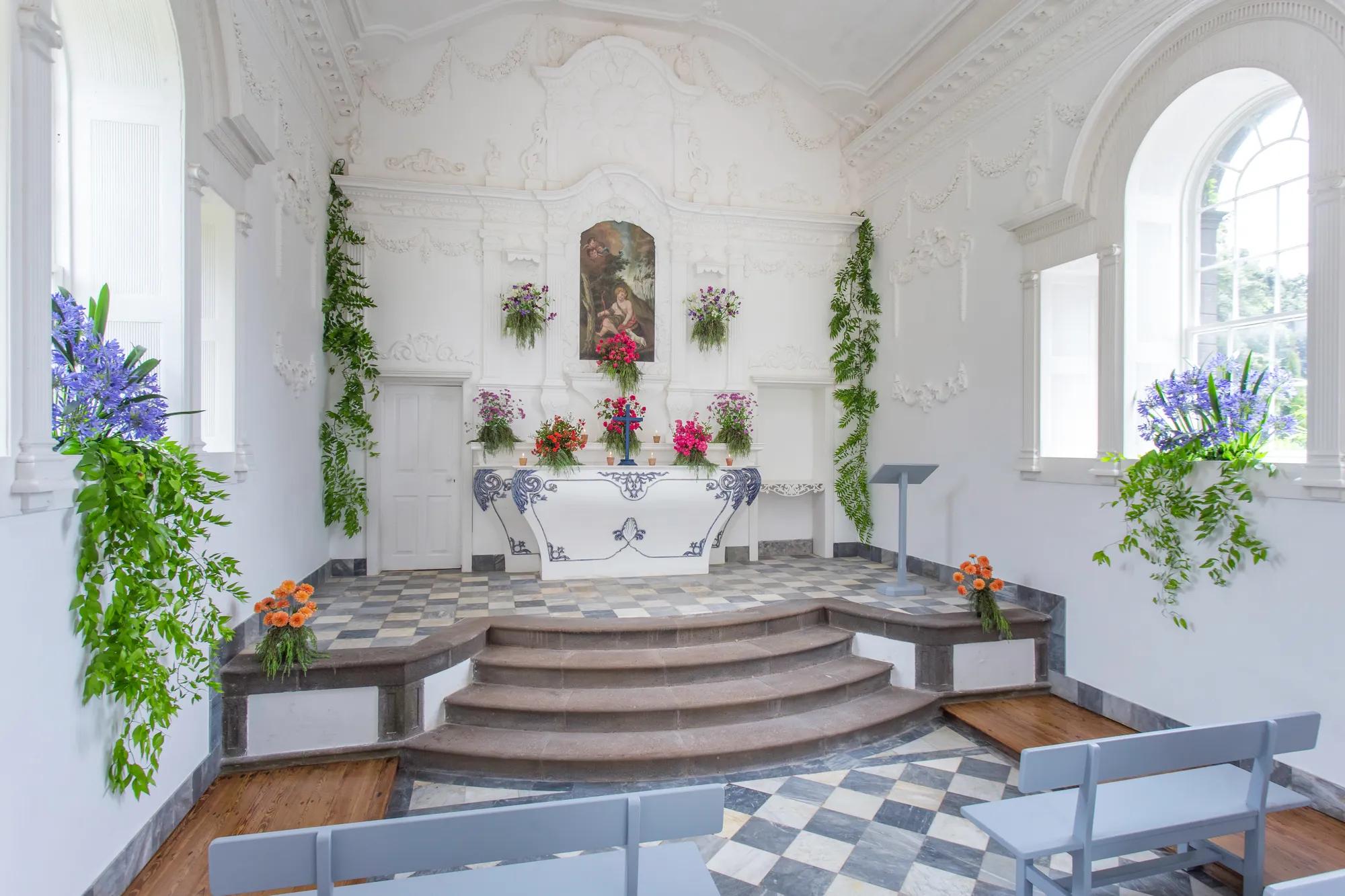Sunlight streams through arched windows into this intimate chapel, where ornate white plasterwork frames vibrant floral arrangements and a peaceful altar painting.