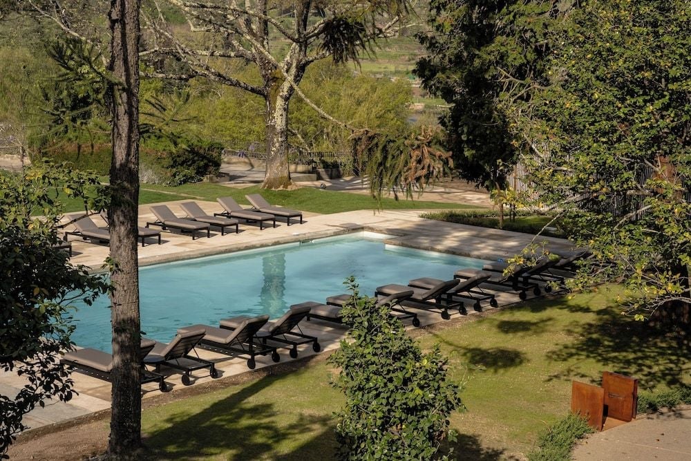 Dappled sunlight filters through ancient trees onto the serene swimming pool, where sleek loungers invite lazy afternoons in this verdant Portuguese retreat.