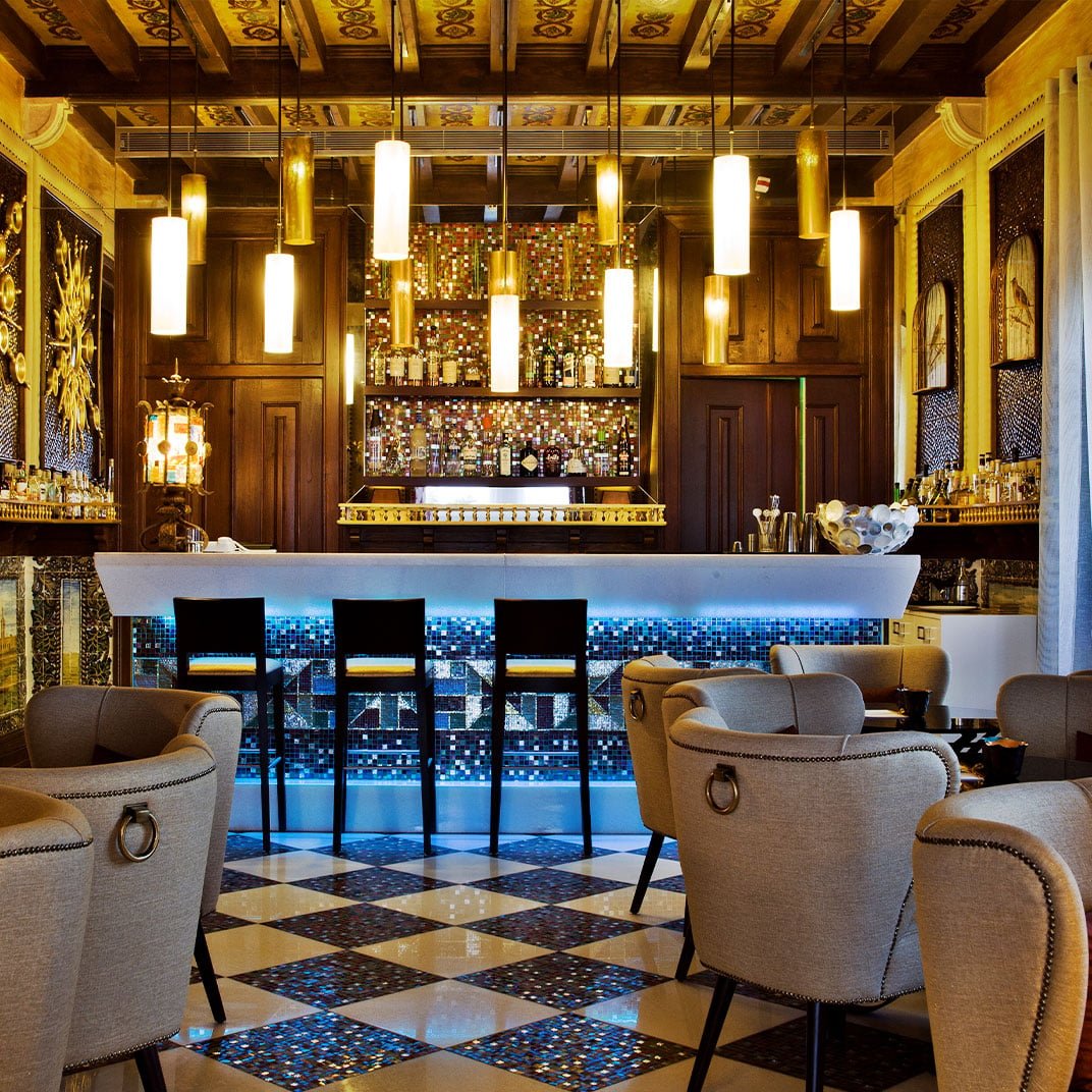 Ornate ceiling frescoes and mosaic tile work create an opulent backdrop where cylindrical pendant lights cast golden hues over plush armchairs and polished marble floors.