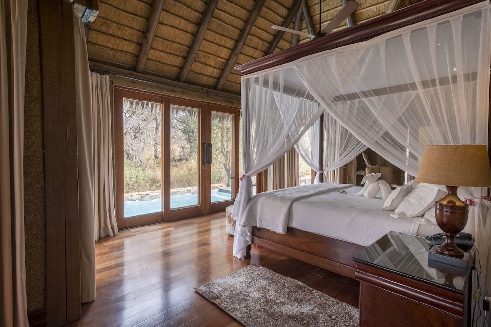 Gossamer mosquito netting drapes the four-poster bed while thatched roofing and polished hardwood floors create an elevated safari sanctuary overlooking the African wilderness.