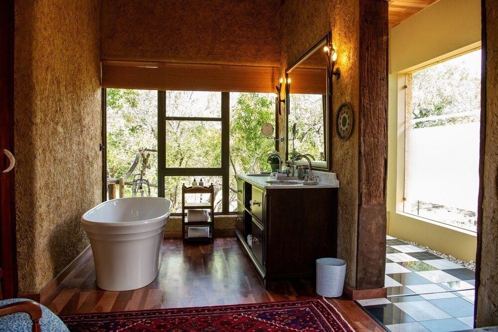 Raw earth walls frame a sculptural soaking tub in this open-concept suite, where African sunlight streams through floor-to-ceiling windows overlooking the wilderness.