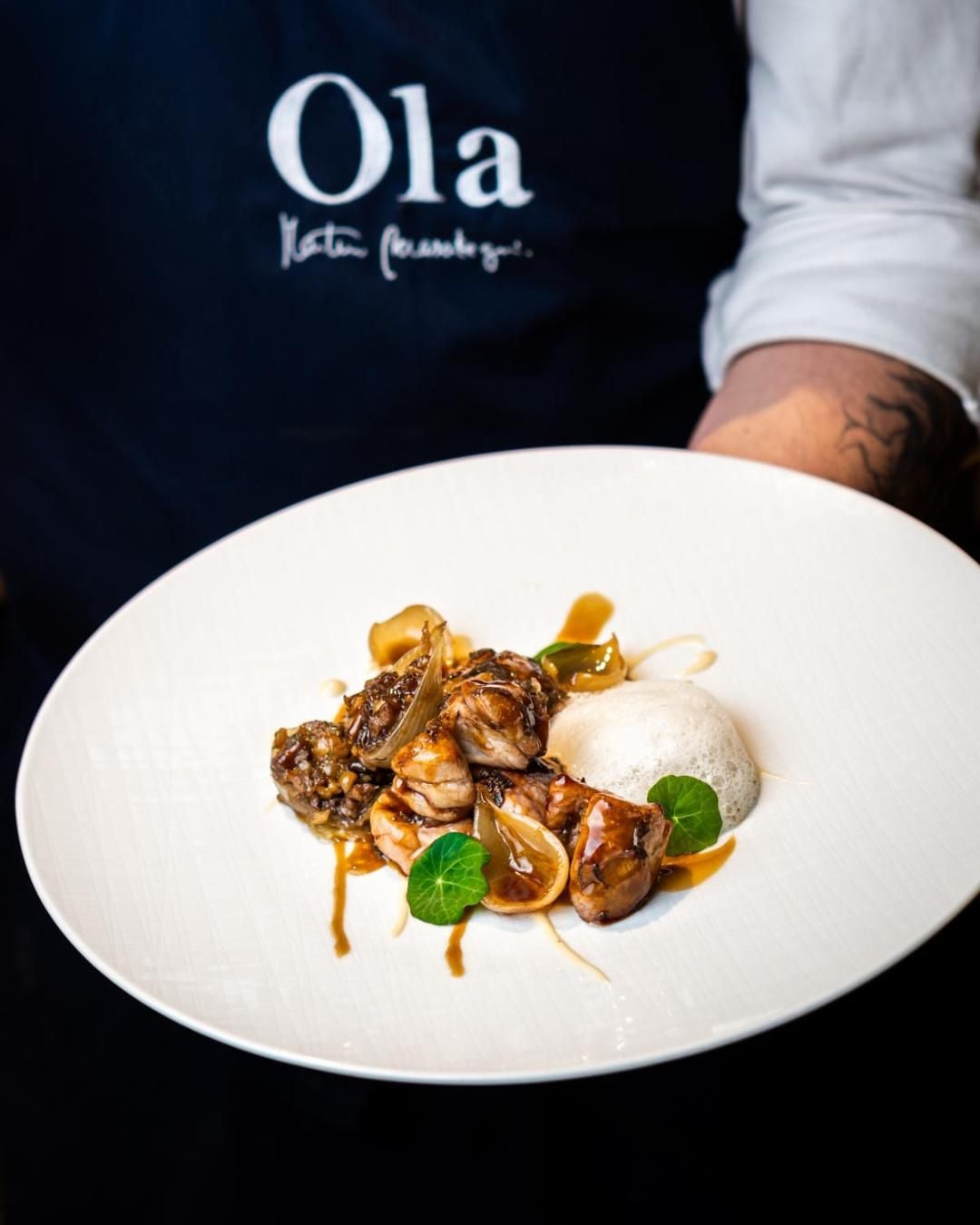 Ola's kitchen artistry shines through seared octopus paired with silky foam and caramelized onions, each element precisely plated on pristine white porcelain.