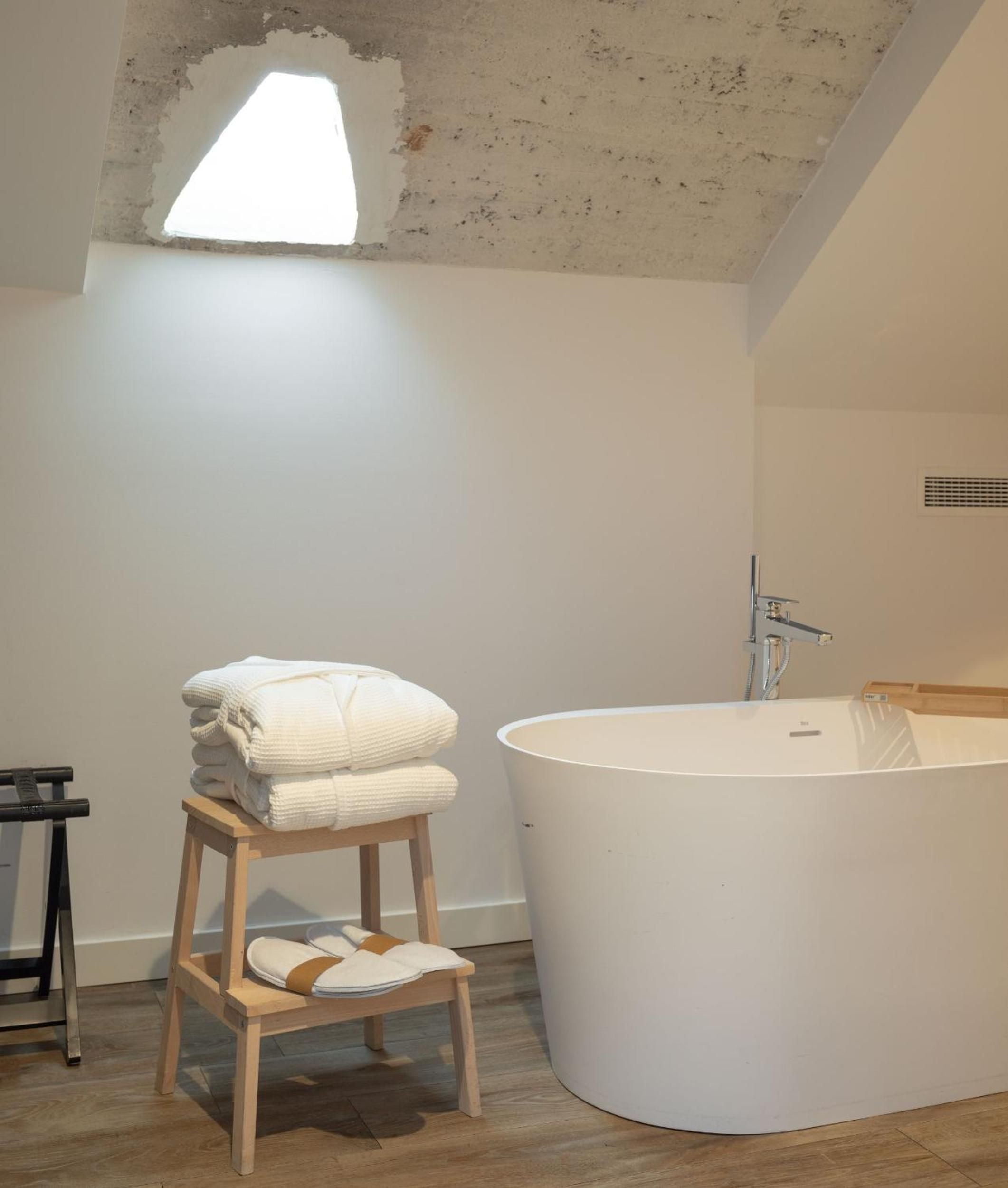 Raw concrete meets refined luxury in this attic bathroom, where natural light filters through weathered stone and fluffy robes await beside the sculptural soaking tub.