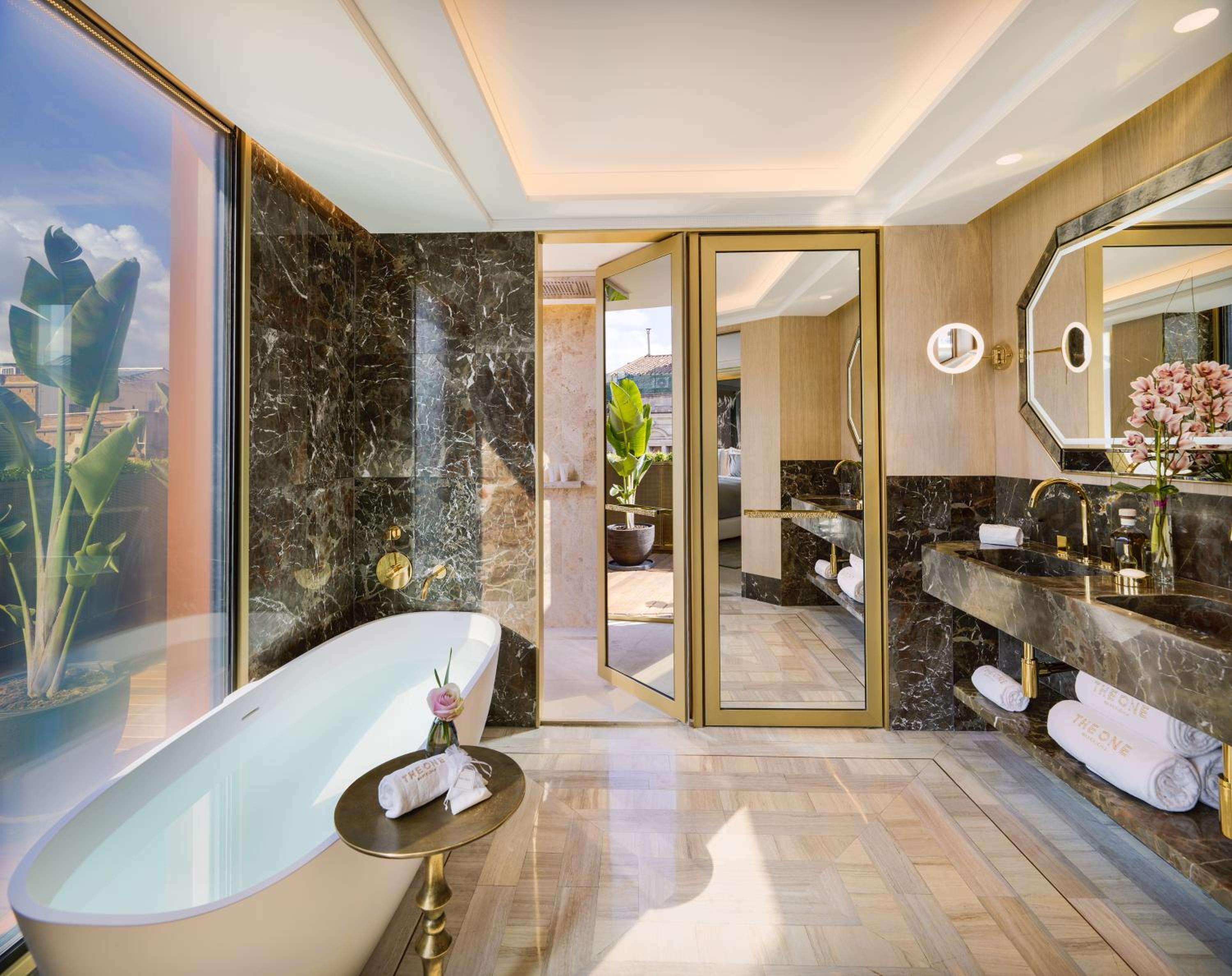 Sunlight streams through floor-to-ceiling windows into this marble sanctuary, where a freestanding soaking tub overlooks verdant tropical foliage beyond gleaming brass fixtures.