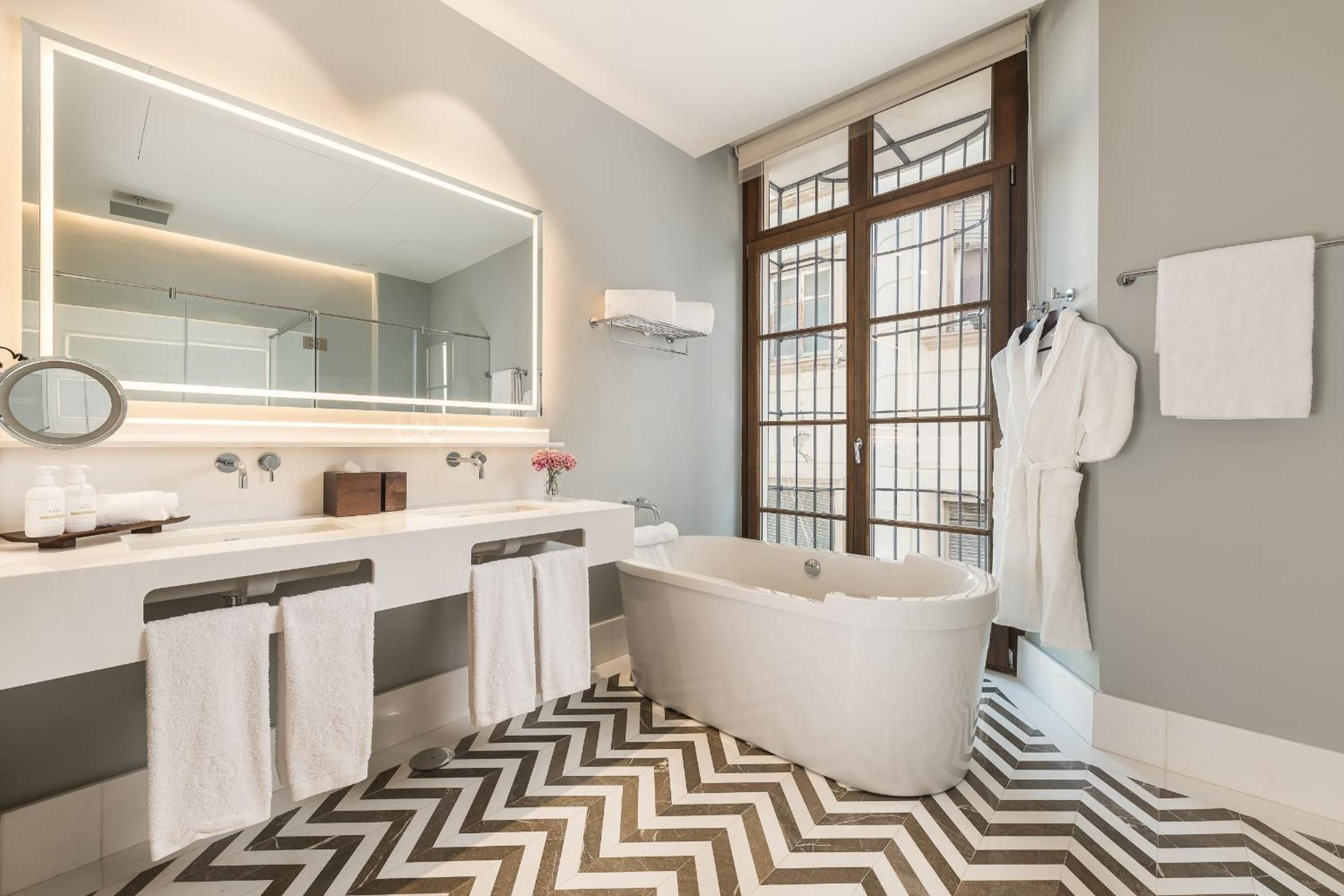Chevron marble floors create dramatic geometry beneath the freestanding soaking tub, while backlit mirrors and crisp white robes promise pure indulgence in this sophisticated sanctuary.