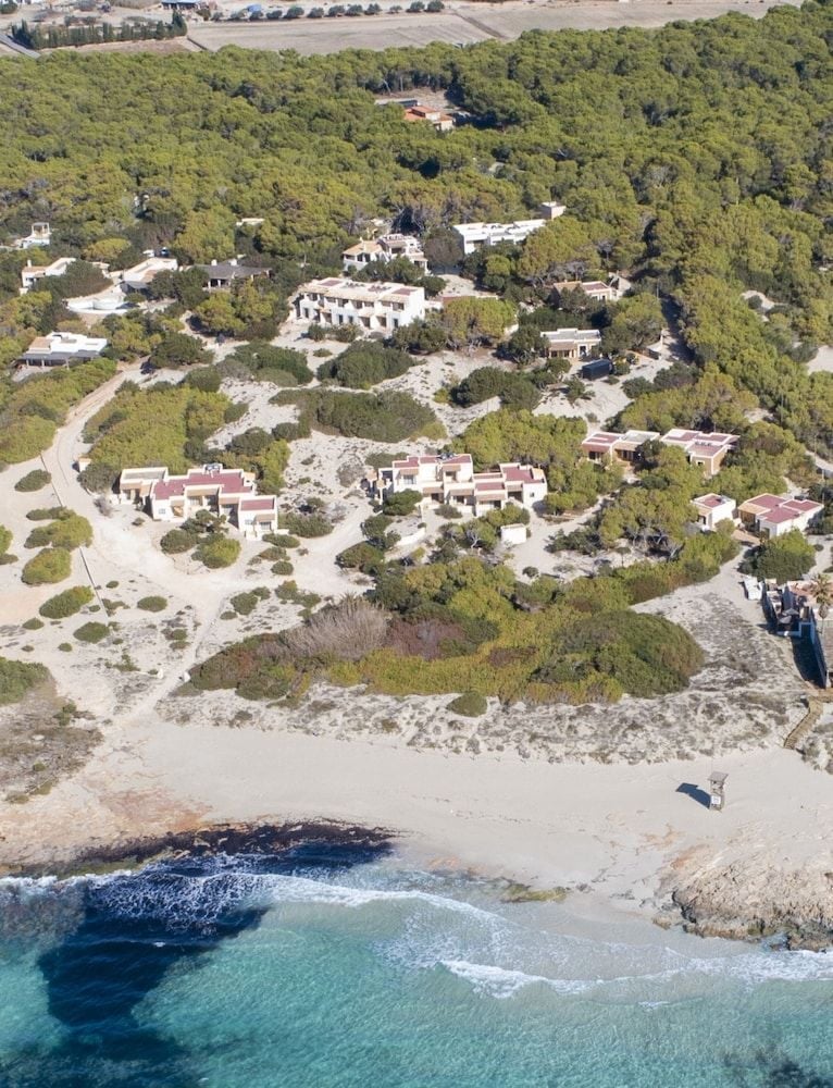 Mediterranean pine forests embrace this secluded coastal retreat, where terracotta-roofed villas nestle between emerald waters and untamed Balearic wilderness on Formentera's pristine shores.