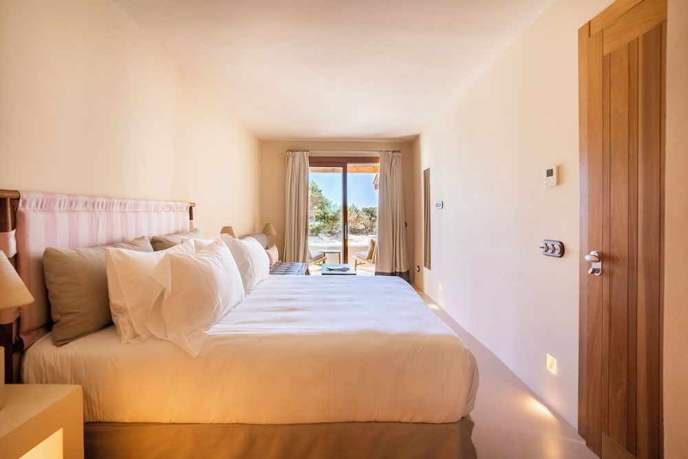 Crisp white linens and warm wood accents create a serene sanctuary, while French doors frame glimpses of Formentera's wild coastline beyond.