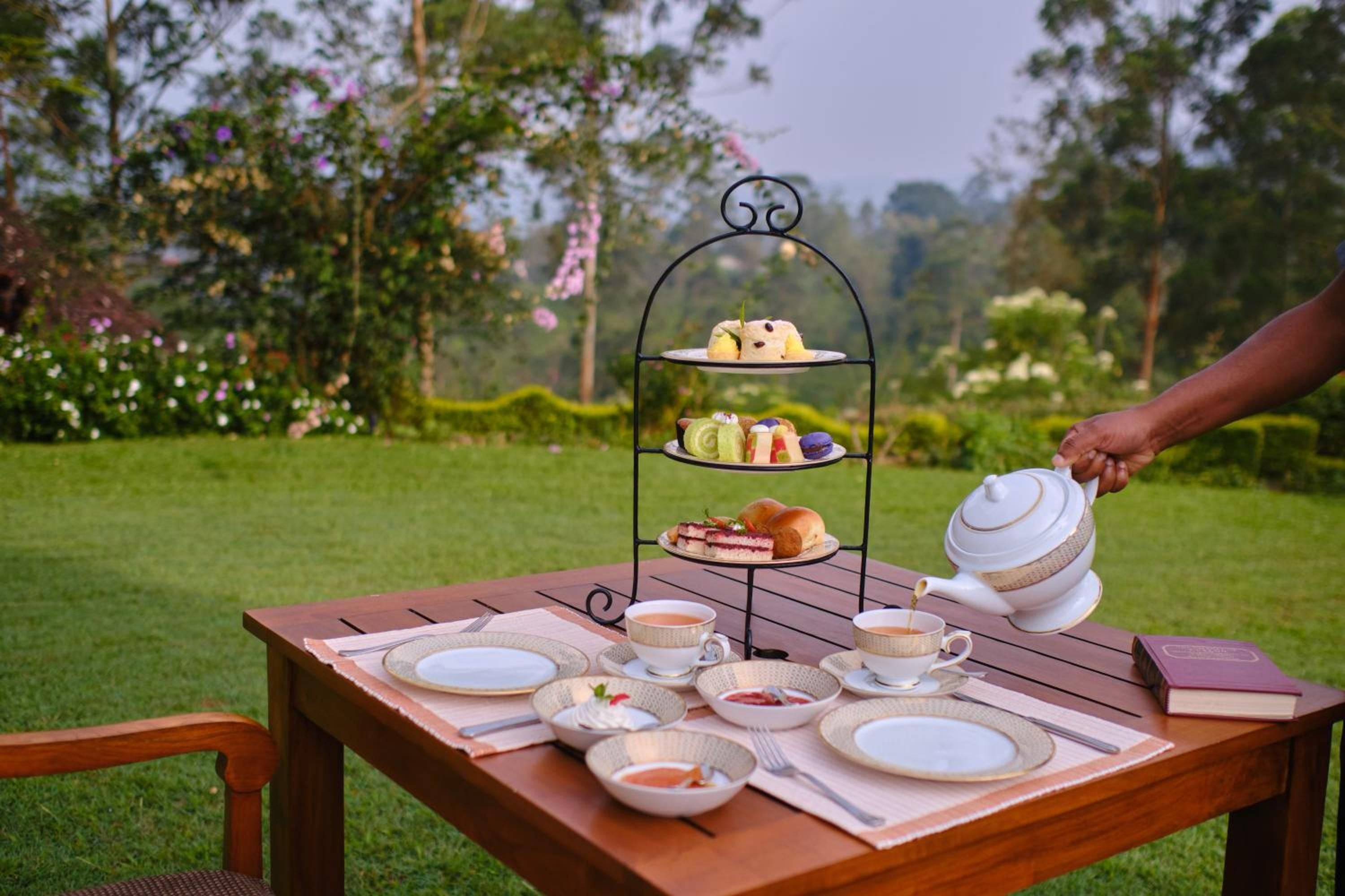 Afternoon tea unfolds on polished teak beneath towering trees, where delicate petit fours and steaming Ceylon tea create moments of pure indulgence in the estate gardens.
