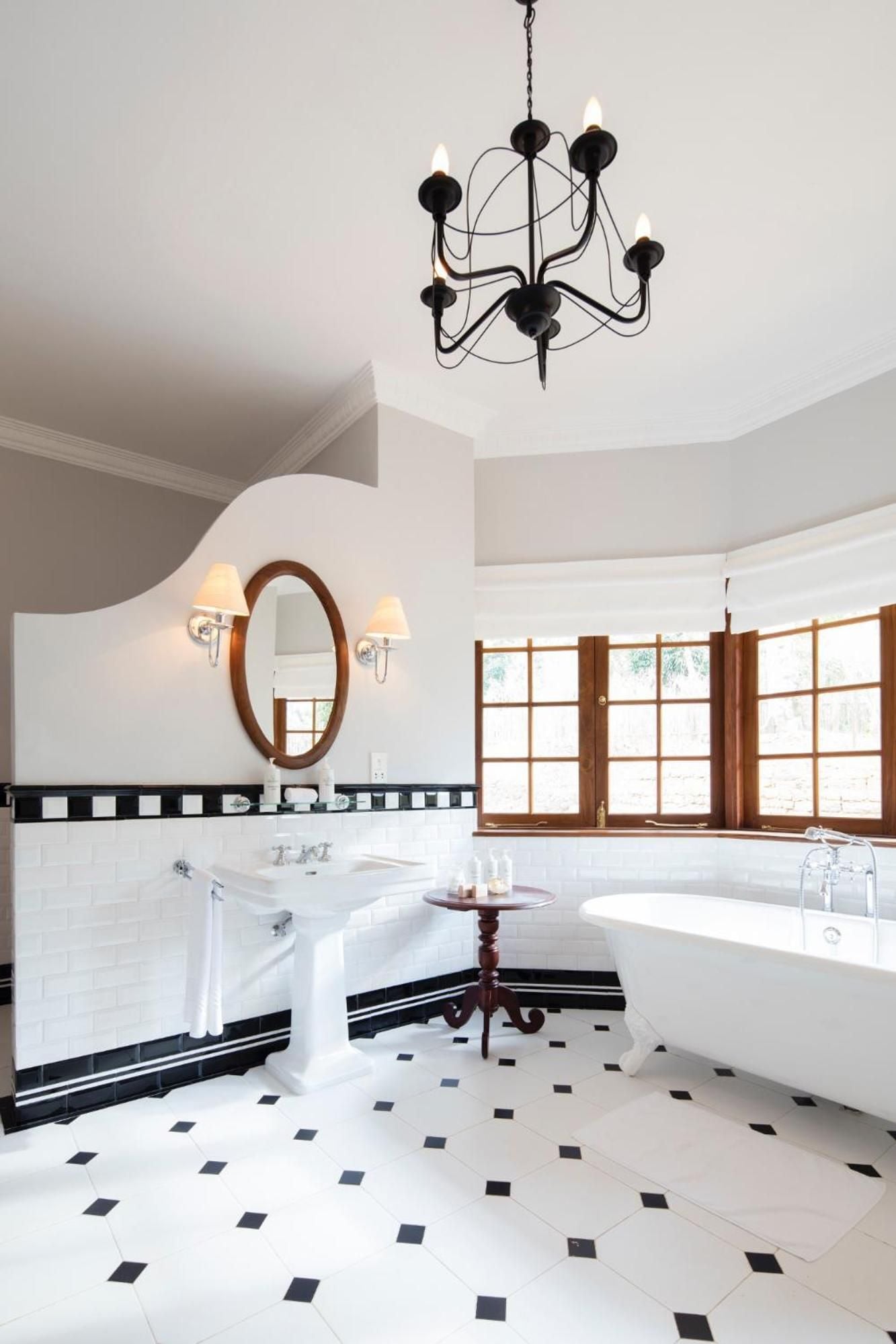 Black and white checkered floors and vintage brass fixtures create old-world elegance in this palatial suite bathroom, where colonial charm meets modern luxury beneath soaring ceilings.
