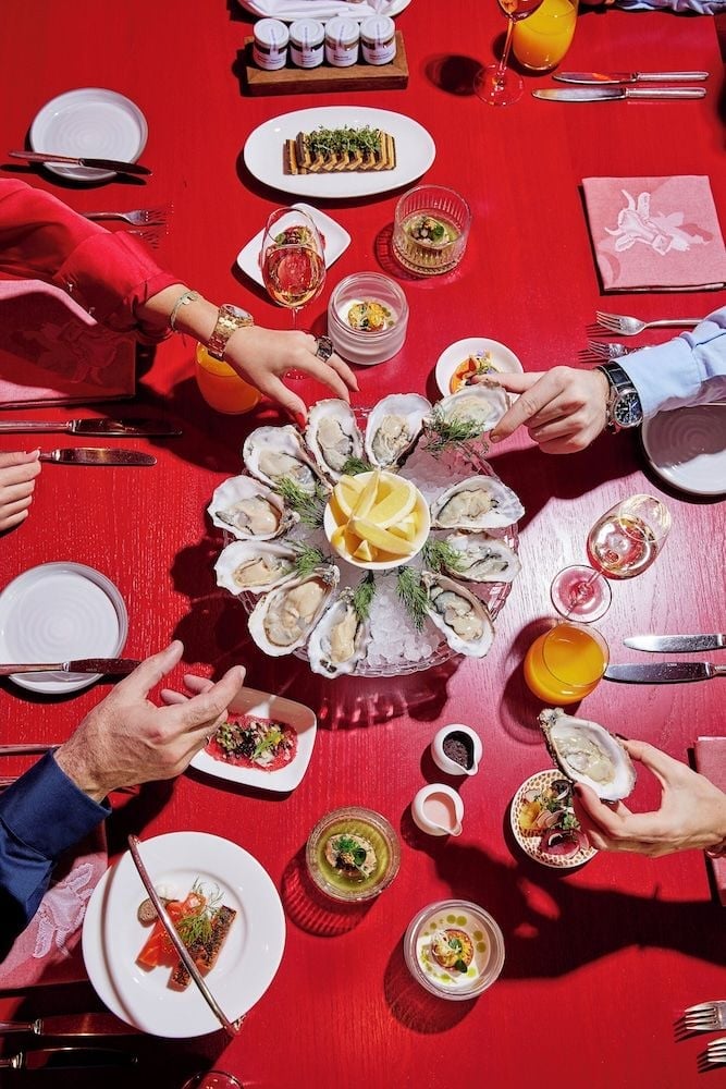 Fresh oysters arranged on crushed ice anchor an elegant spread where hands reach for delicate bites across the crimson tablecloth at this refined Alpine dining experience.