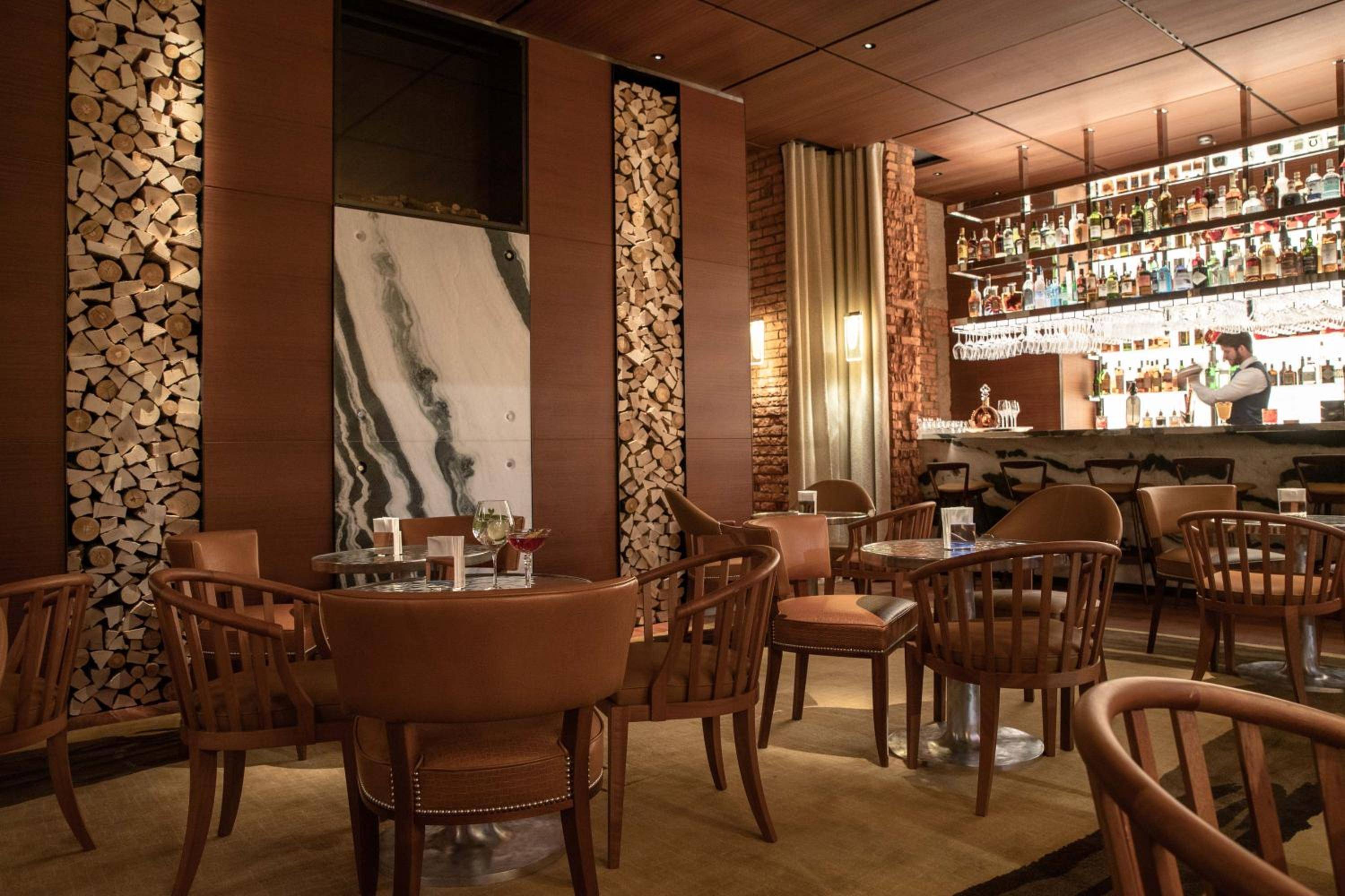 Warm wood textures and stacked logs create intimate ambiance in the hotel's sophisticated bar, where craft cocktails meet alpine-inspired design details.