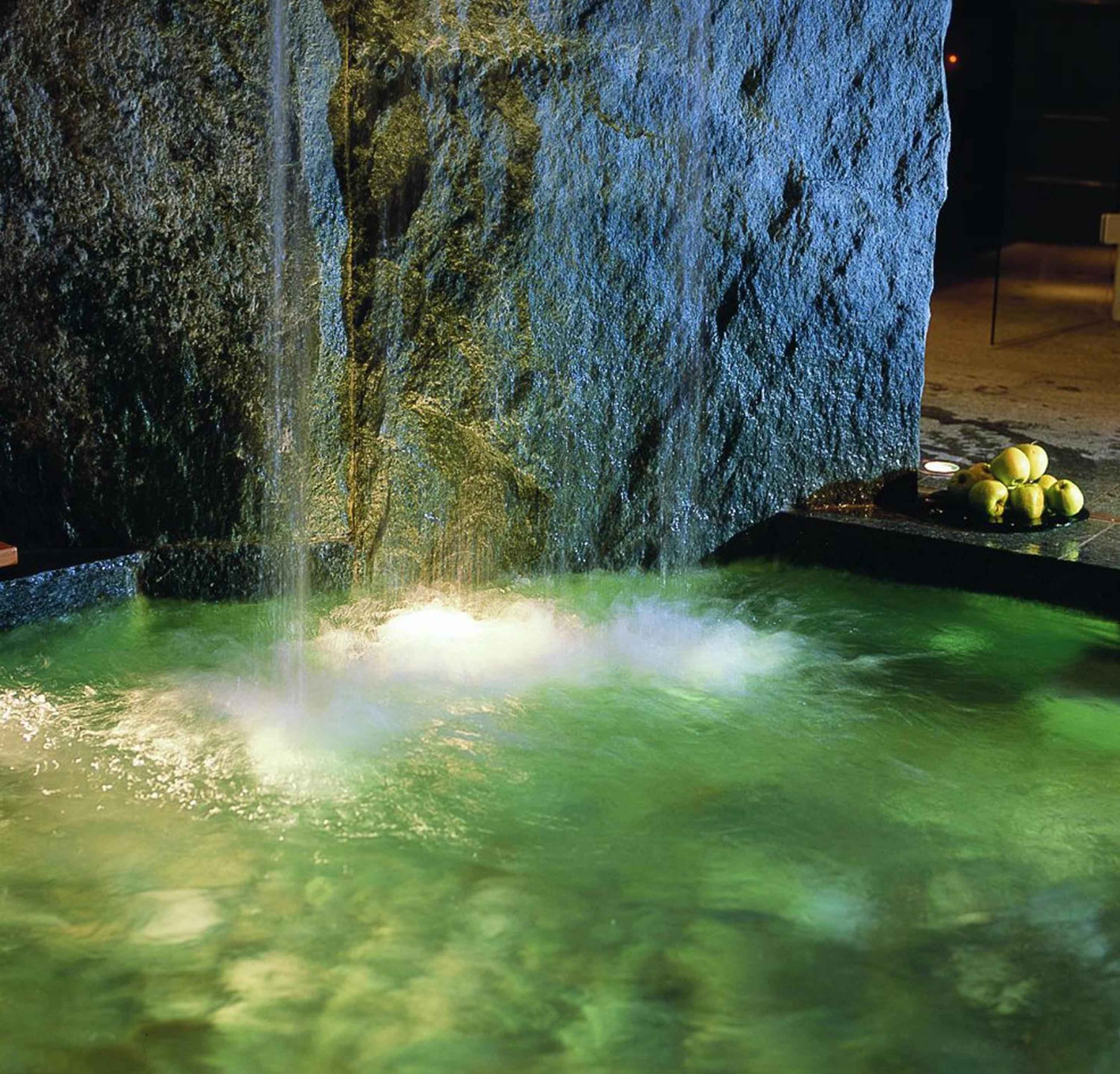 Steam rises from jade-green thermal waters in this intimate grotto spa, where ancient stone walls glisten with moisture and golden light creates an otherworldly sanctuary.