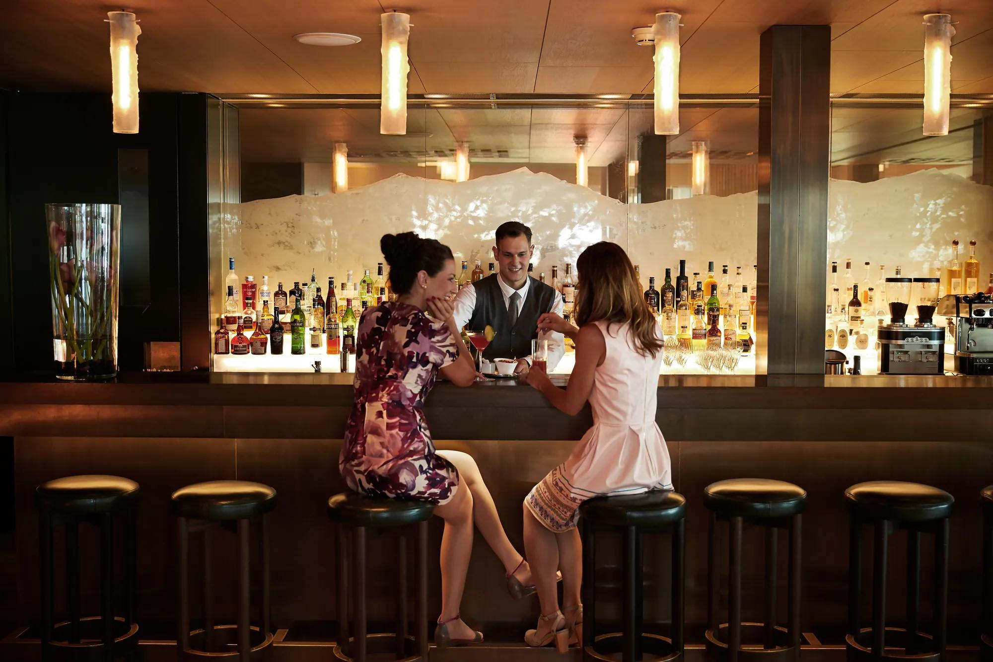 Pendant lights cast golden warmth over polished marble as guests savor expertly crafted cocktails, the backlit bottle display creating an intimate glow for evening conversations.