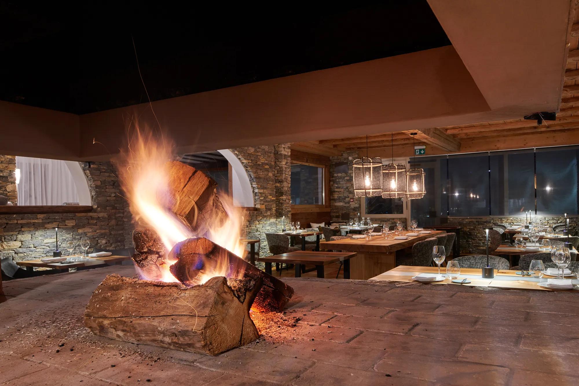 Crackling logs cast dancing shadows across stone walls while diners gather around candlelit tables, creating an intimate alpine dining experience that feels authentically rustic yet refined.