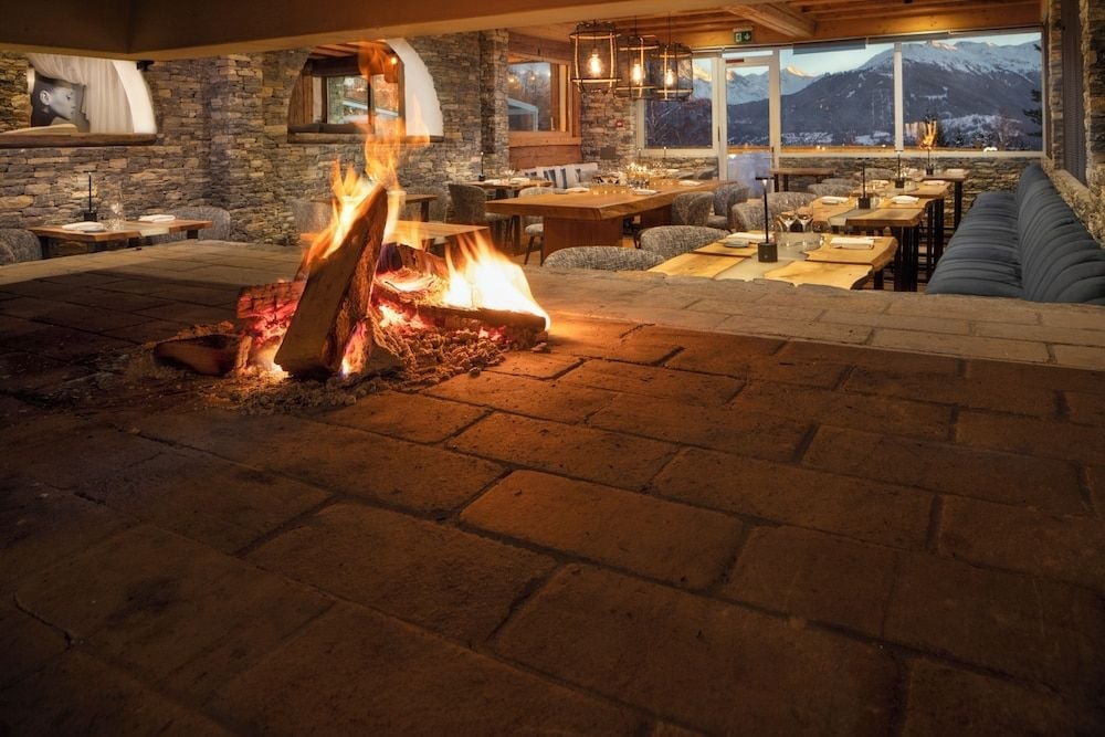 Crackling flames cast golden light across stone floors while snow-capped peaks frame intimate dining tables in this mountain lodge restaurant.