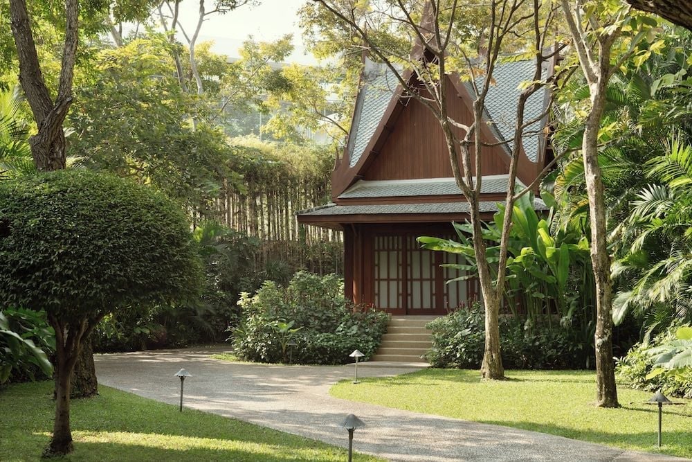 Weathered teak pavilions emerge from tropical gardens like ancient temples, their steep-pitched roofs catching dappled sunlight through towering palms and flowering shrubs.