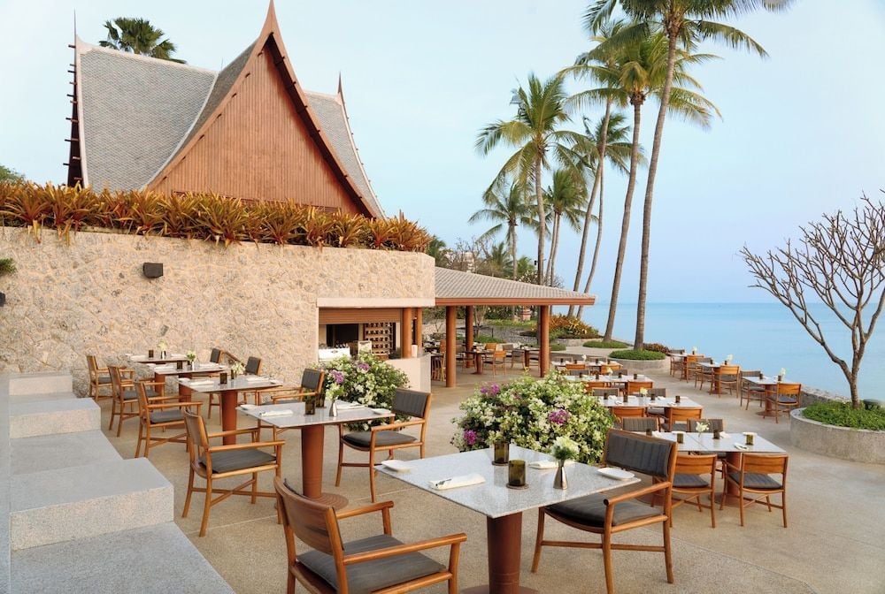 Teakwood tables dot the oceanfront terrace where Thai temple spires frame alfresco dining, palm trees swaying against azure horizons at this legendary wellness retreat.