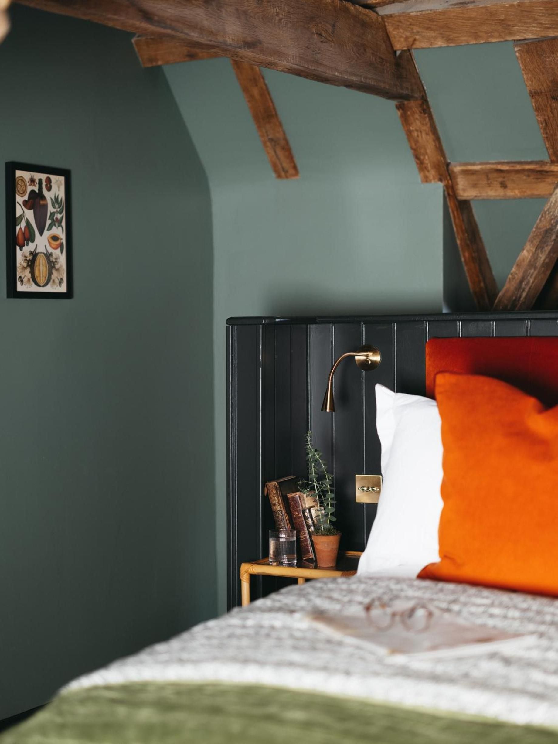 Exposed timber beams frame sage-green walls in this intimate bedroom, where brass reading lights and burnt orange linens create a cozy countryside retreat.
