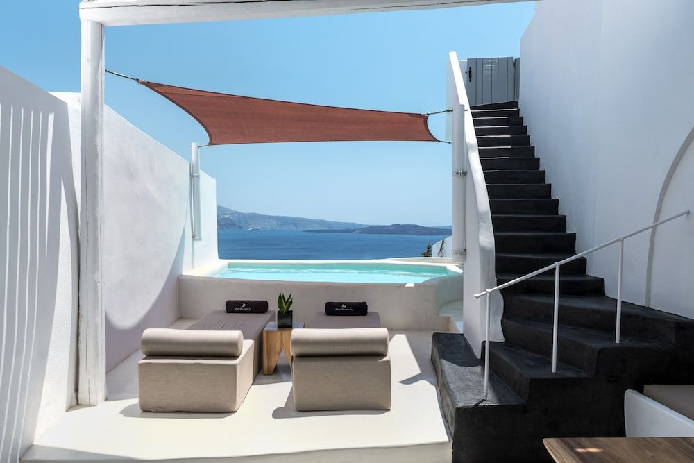 Cream loungers frame an infinity pool that bleeds into Santorini's endless blue, where terracotta sail cloth filters Mediterranean light across pristine white terraces.
