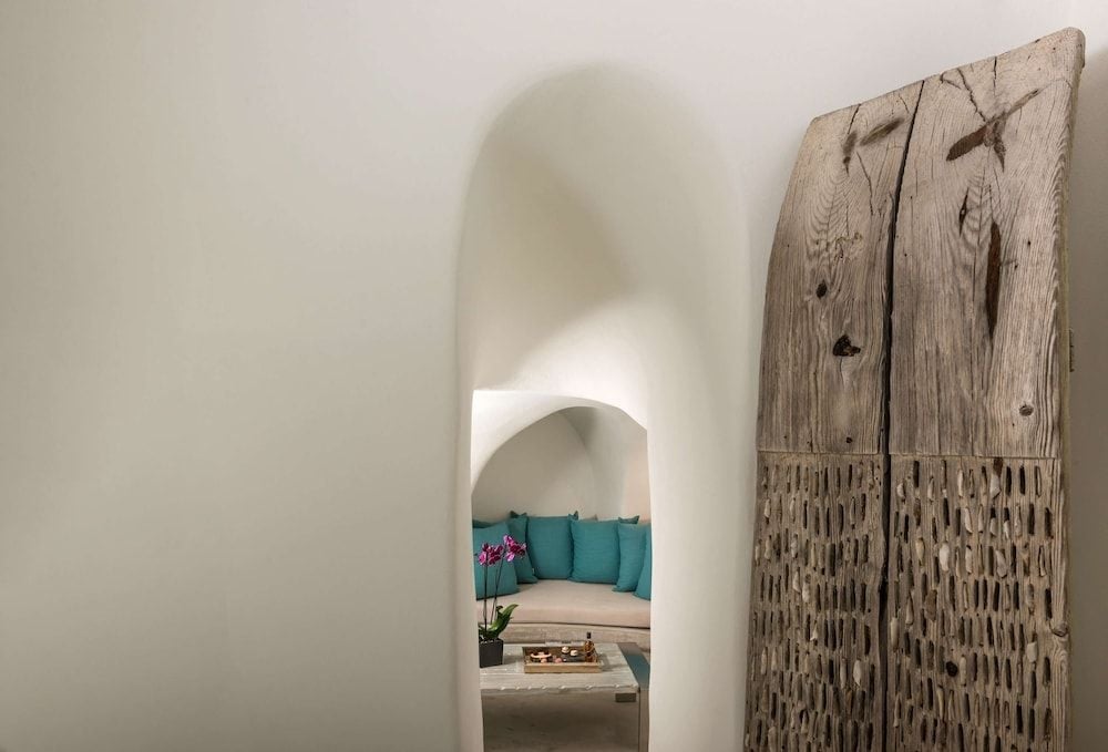 Weathered driftwood doors frame an arched passage where turquoise cushions beckon from sun-drenched seating areas, embodying Santorini's seamless blend of rustic charm and refined comfort.