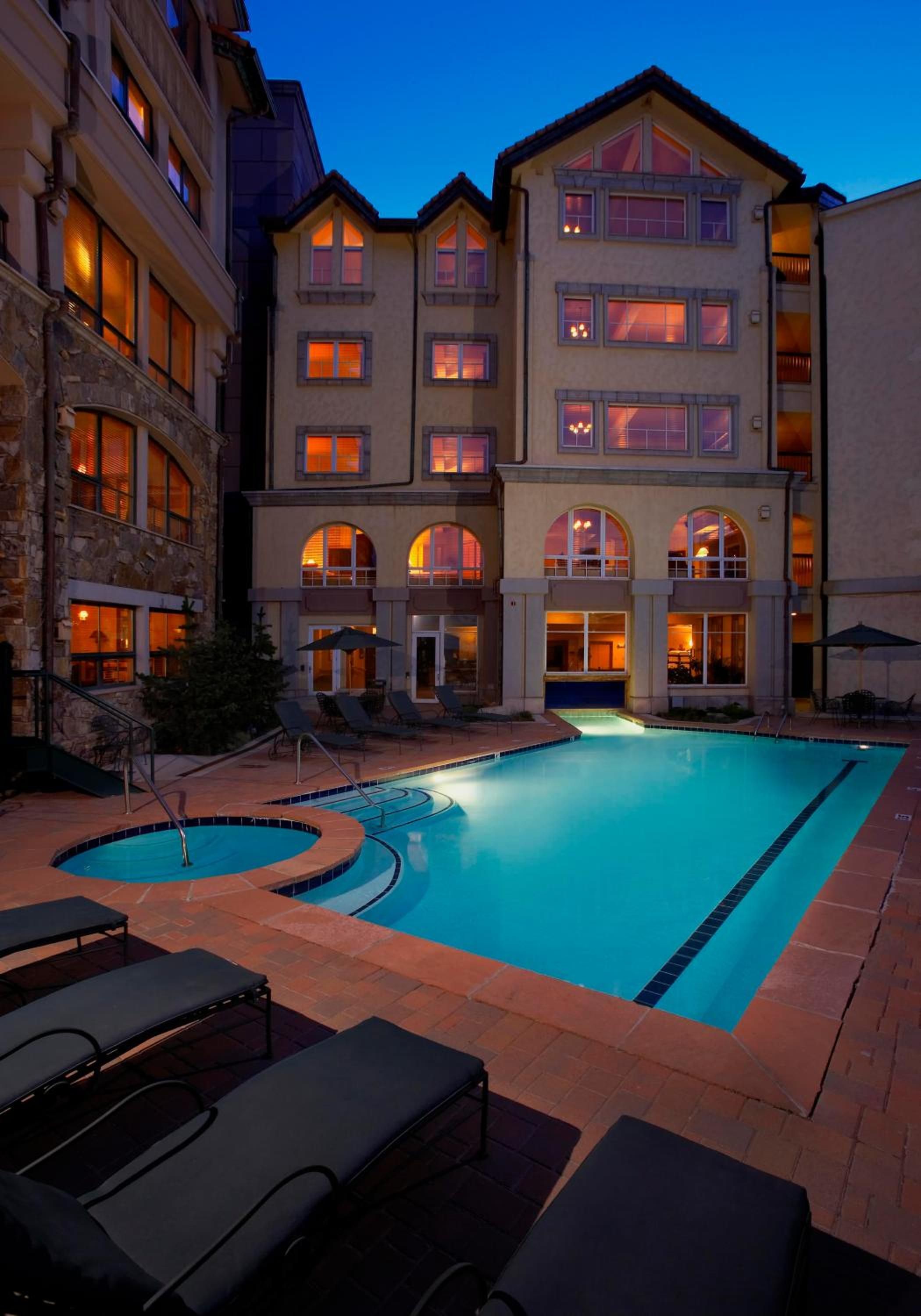 Evening light transforms the heated pool into a turquoise jewel, while warm amber glows spill from arched windows across the alpine-inspired lodge's distinctive stone and stucco facade.