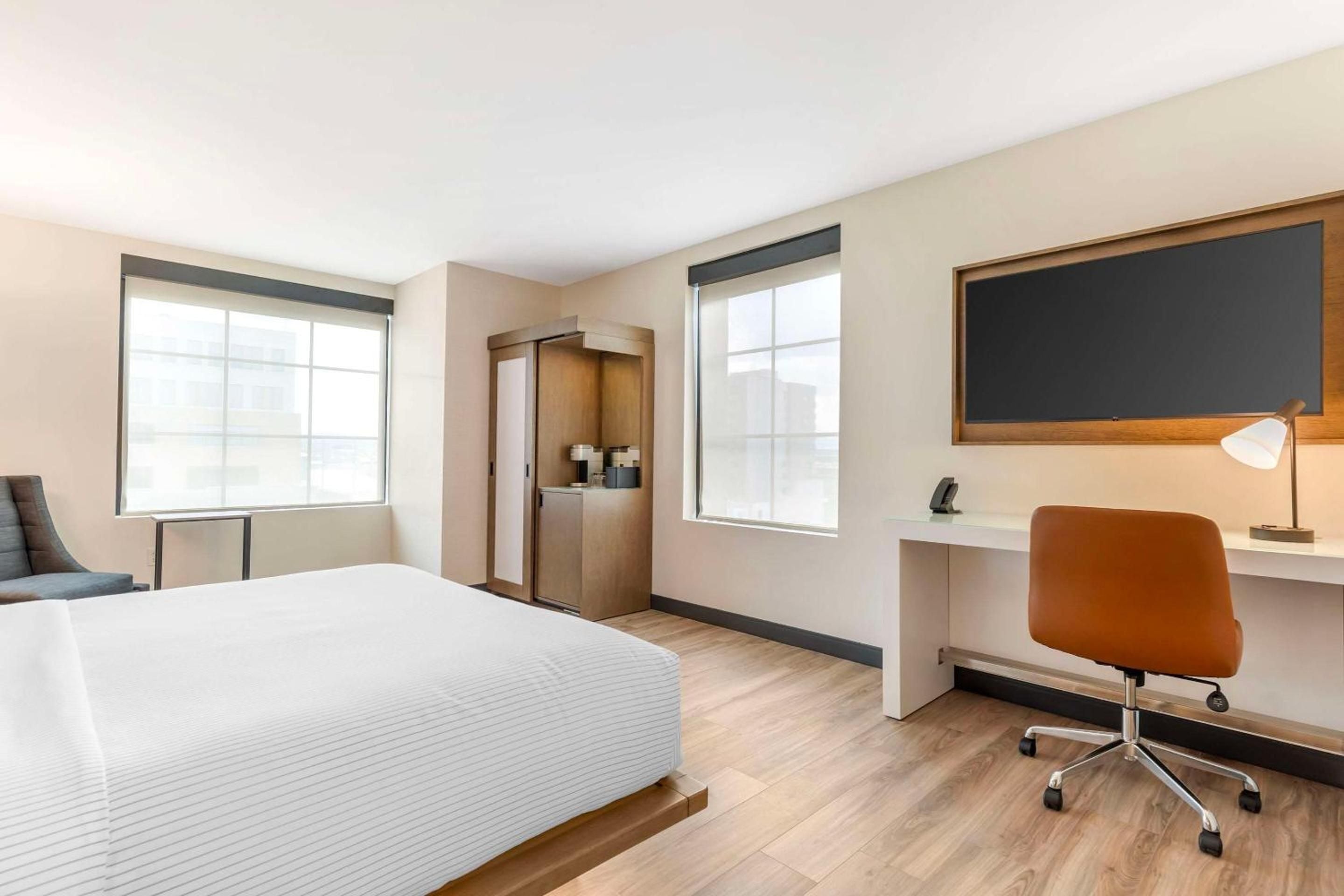 Modern guest rooms blend warm wood tones with crisp white linens, featuring a sleek workspace and minimalist coffee station beneath large windows overlooking Denver's vibrant RiNo district.