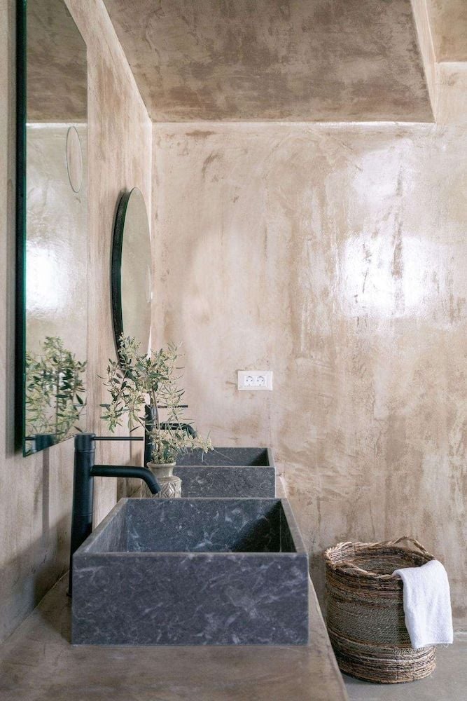Sculptural dark stone basins contrast beautifully against weathered plaster walls, where Mediterranean light filters softly through this serene sanctuary of natural textures and organic forms.